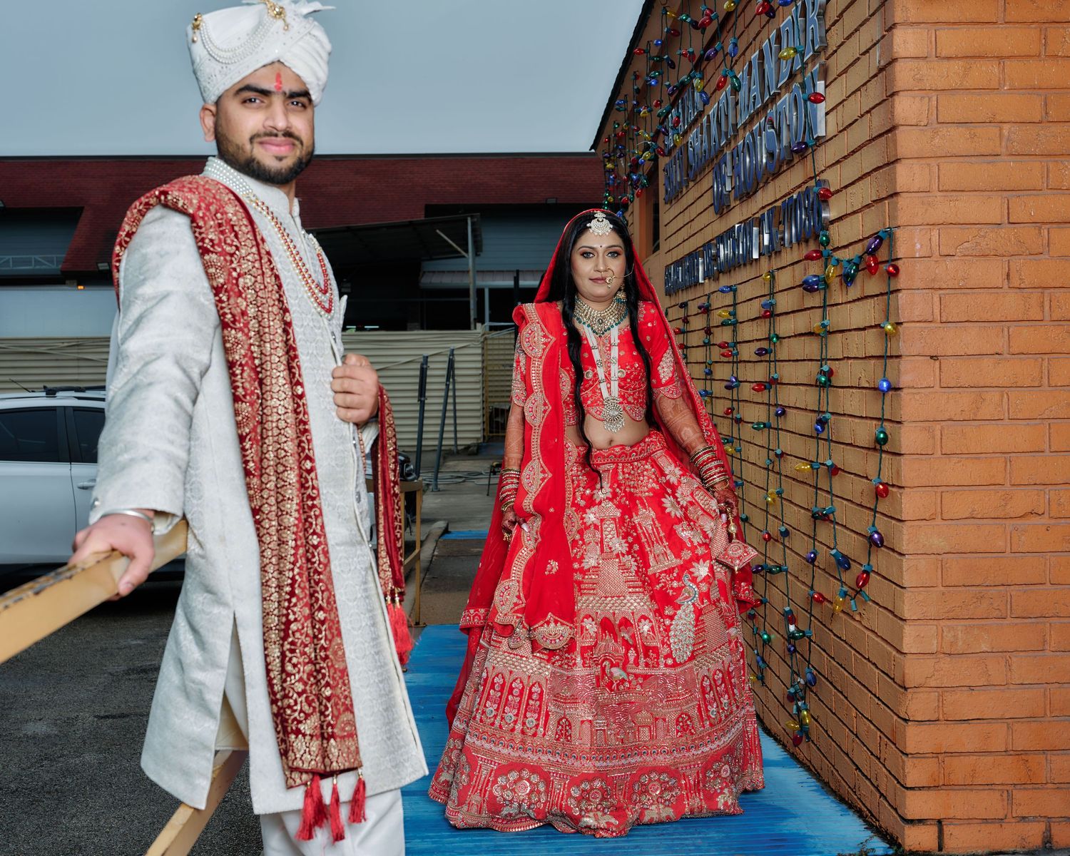 Traditional Indian wedding couple in white and red attire posing against brick wall outdoors.