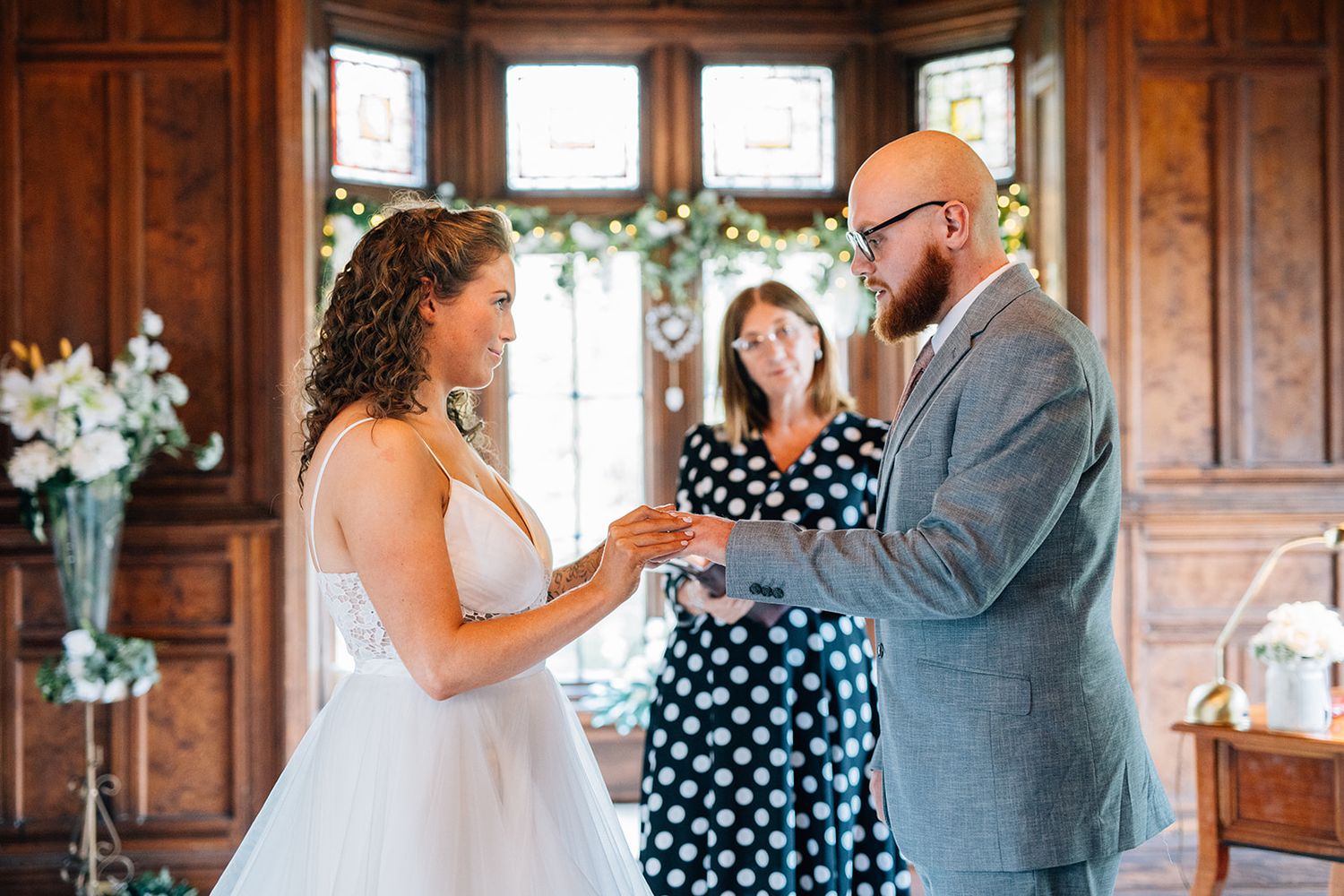 A wedding ceremony takes place in a wood-paneled room with natural light streaming through windows and white floral decorations.