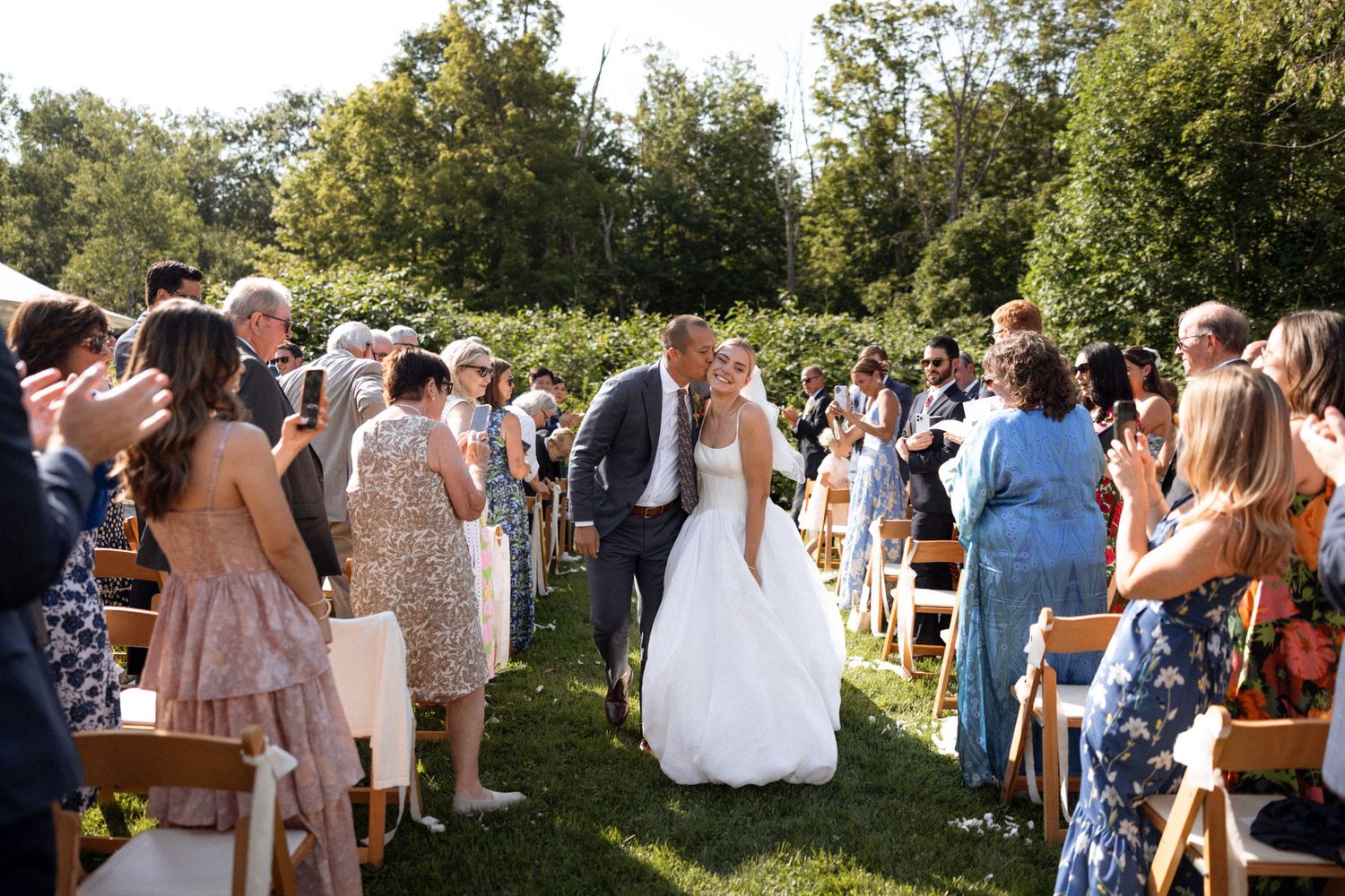 Wedding guests gather outdoors for a ceremony under trees on a sunny day.