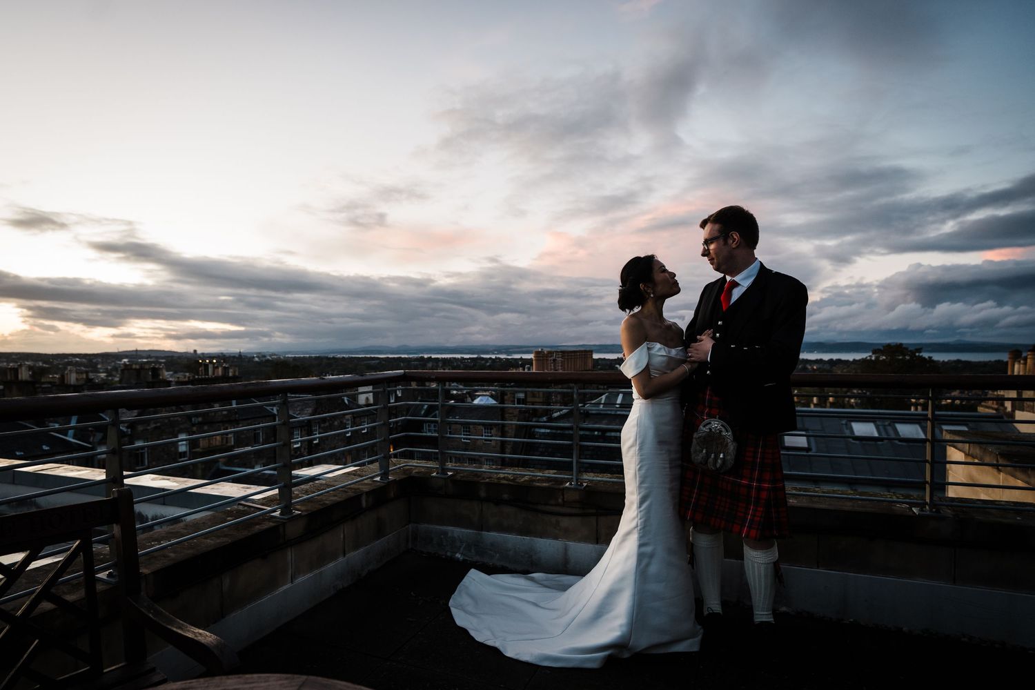 Bride and Groom on rooftop terrace of George Hotel with views over Edinburgh at sunset.  Full length photo, wearing white dress and kilt