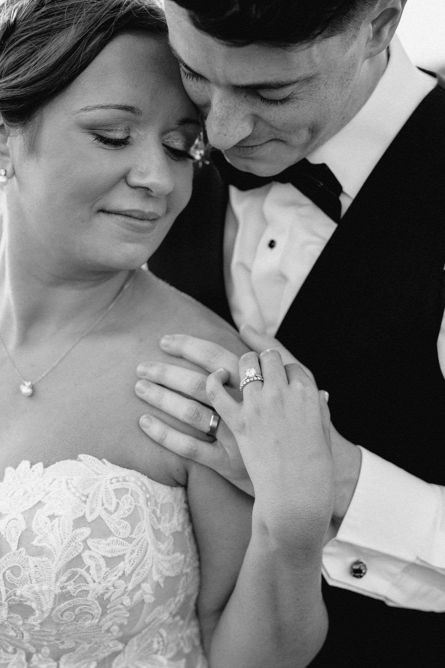 Black and white photo showing close embrace of newlyweds with wedding rings and formal attire.