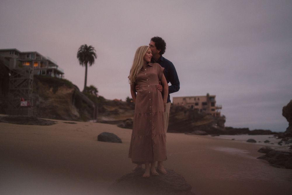 Couple in flowing dresses walk along a dramatic coastal beach during golden hour with cliffs in the background.