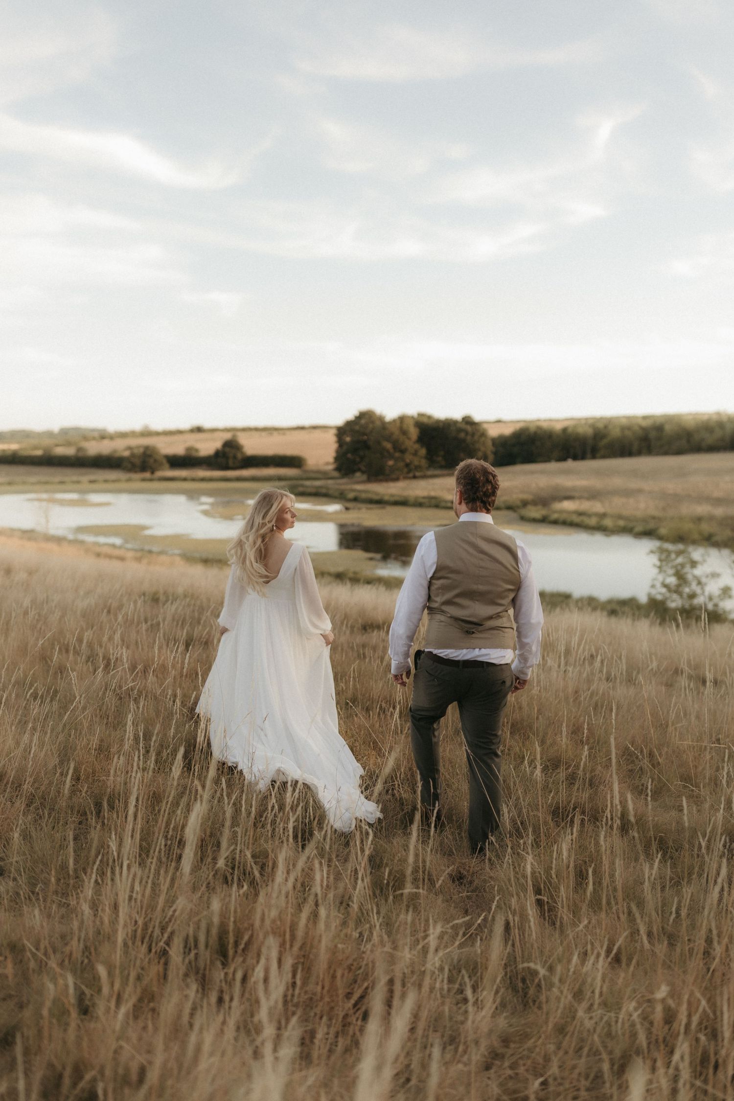 Couple walking together in golden sunlit field during sunset as trees cast long shadows in the background.