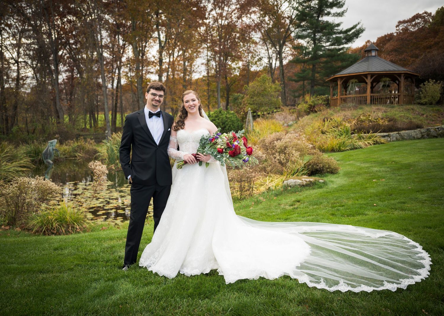 Newlyweds share an intimate moment outdoors with autumn foliage and rustic architecture in background.