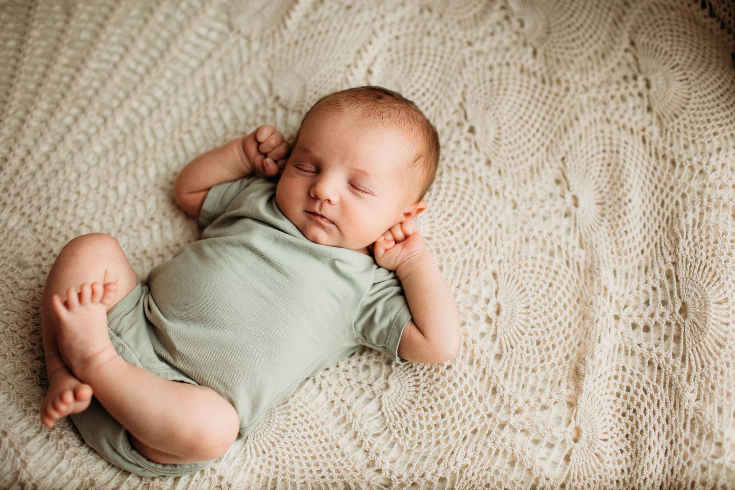 Peacefully sleeping baby wearing mint green onesie lies on cream knitted blanket with bare feet extended.