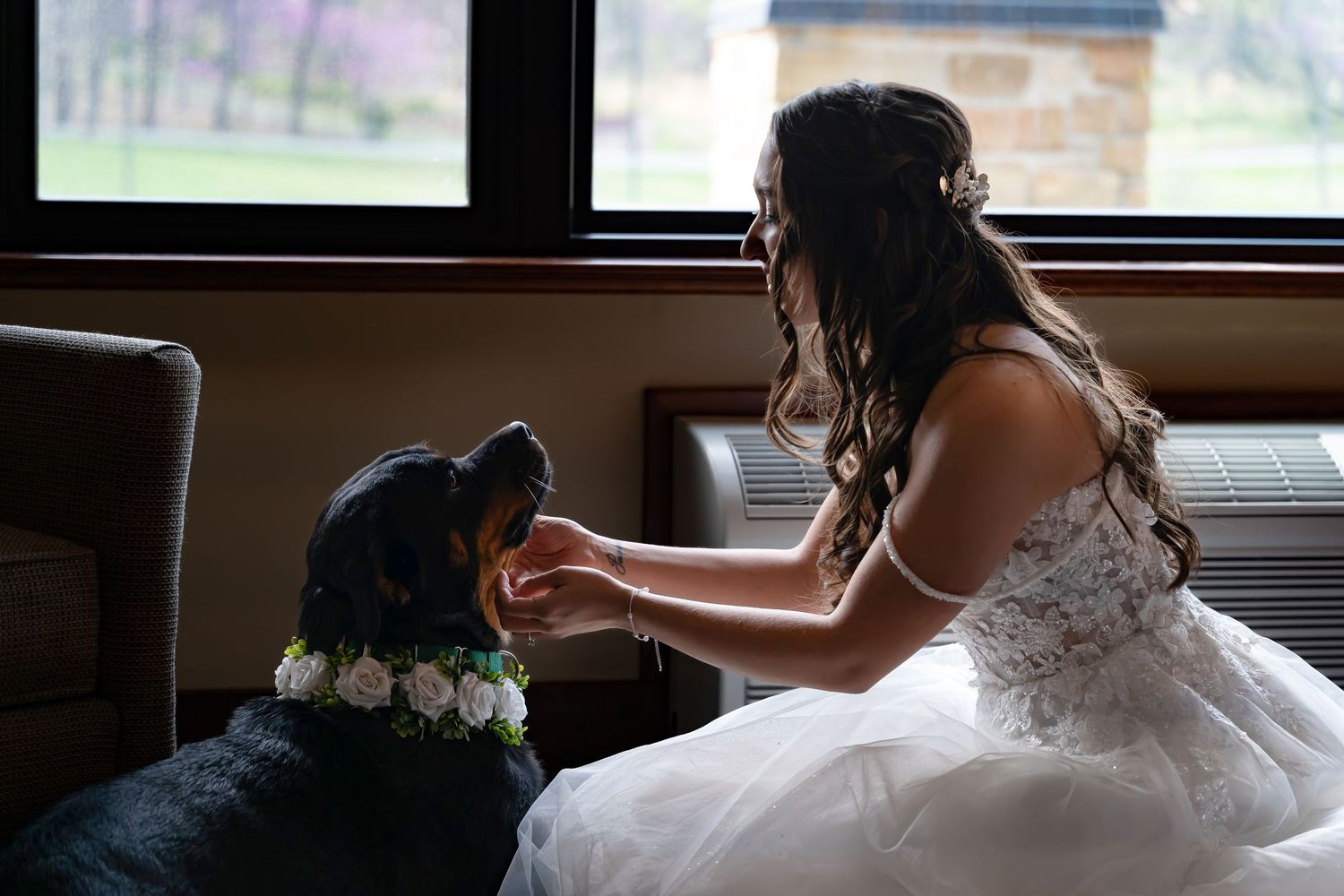 Someone in a white wedding dress sits by a window sharing a tender moment with their black dog.