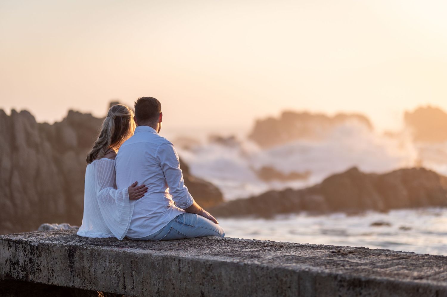 Couple in white sitting on a seaside wall watching waves crash against rocks at golden sunset.