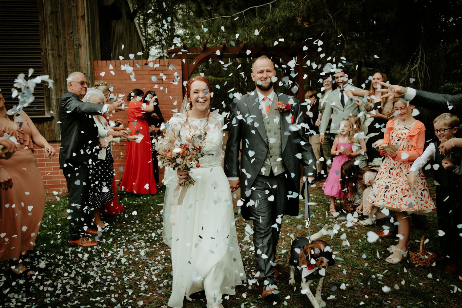 Wedding couple poses among lush greenery and white floral arrangements outdoors.