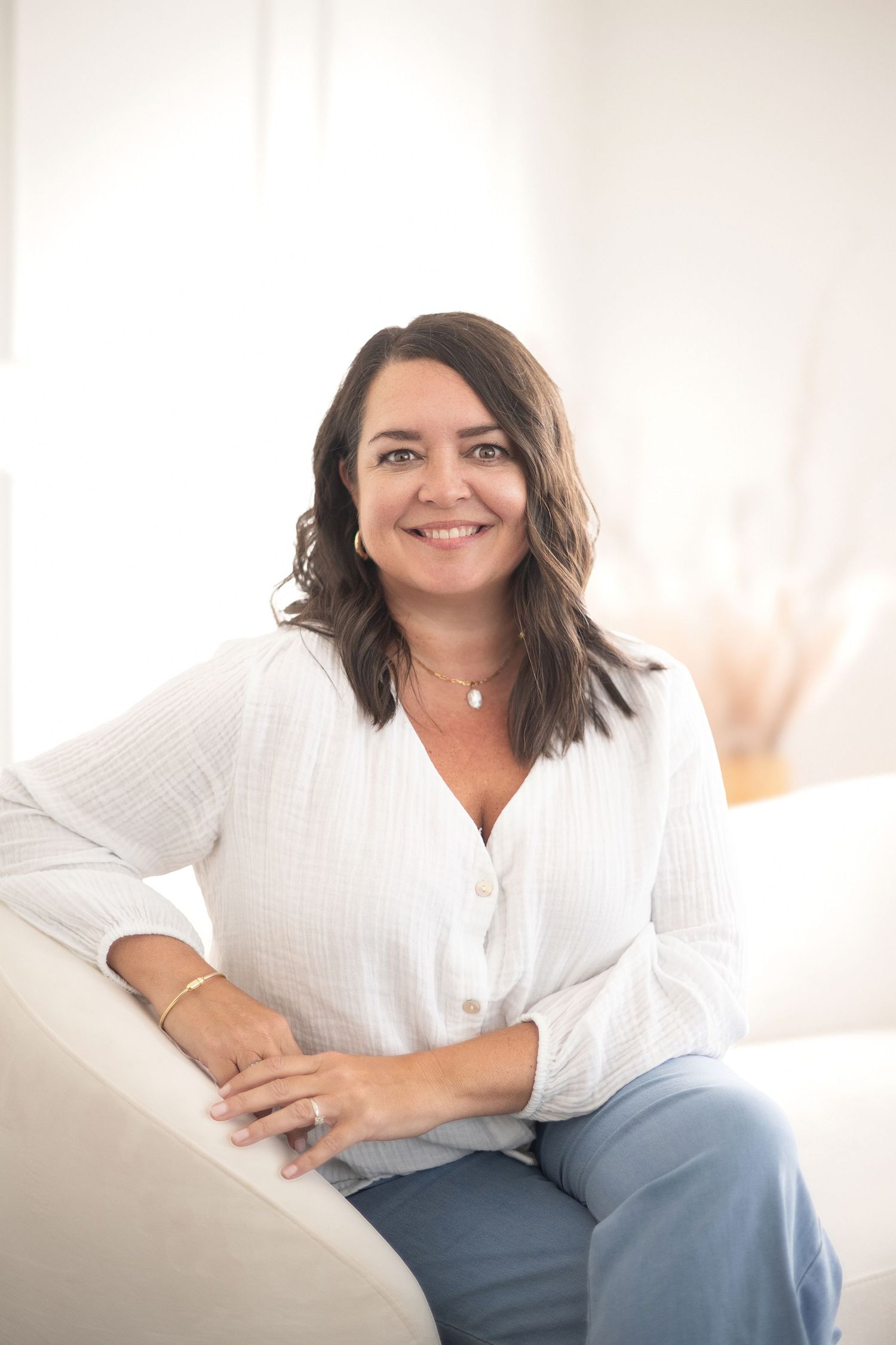 Smiling brunette woman in white linen shirt and jeans sitting relaxed on white sofa in bright room.