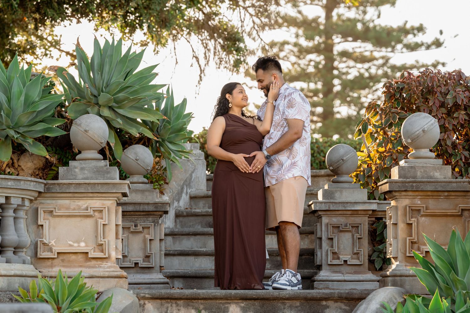 Couple in maternity photo shoot on stone steps surrounded by lush greenery and agave plants at golden hour.