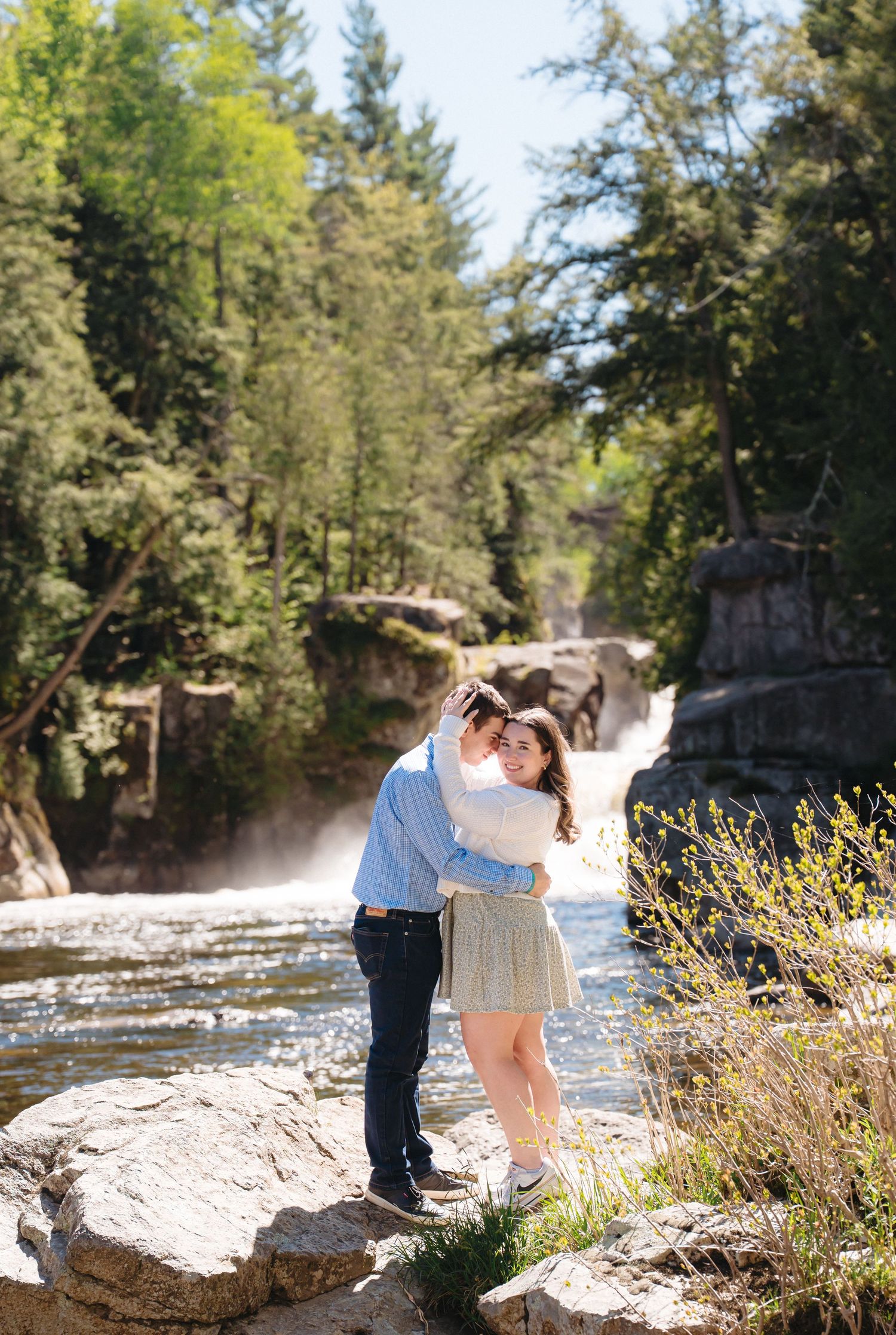 Couple embracing on rocks near a scenic waterfall surrounded by lush green trees on a sunny day.
