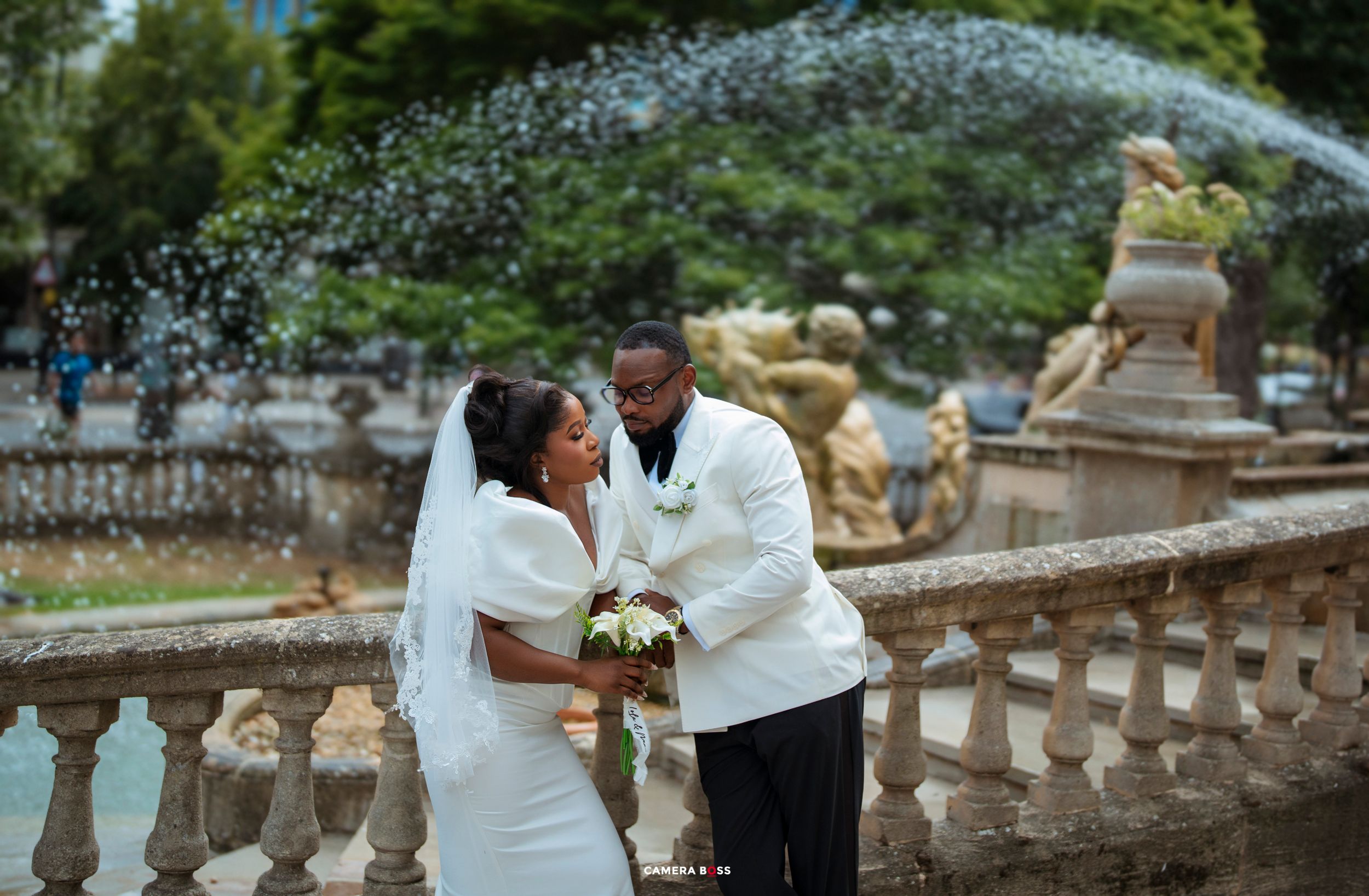 Bride and groom share a romantic moment in front of an ornate fountain, bride holding a bouquet in a white gown.