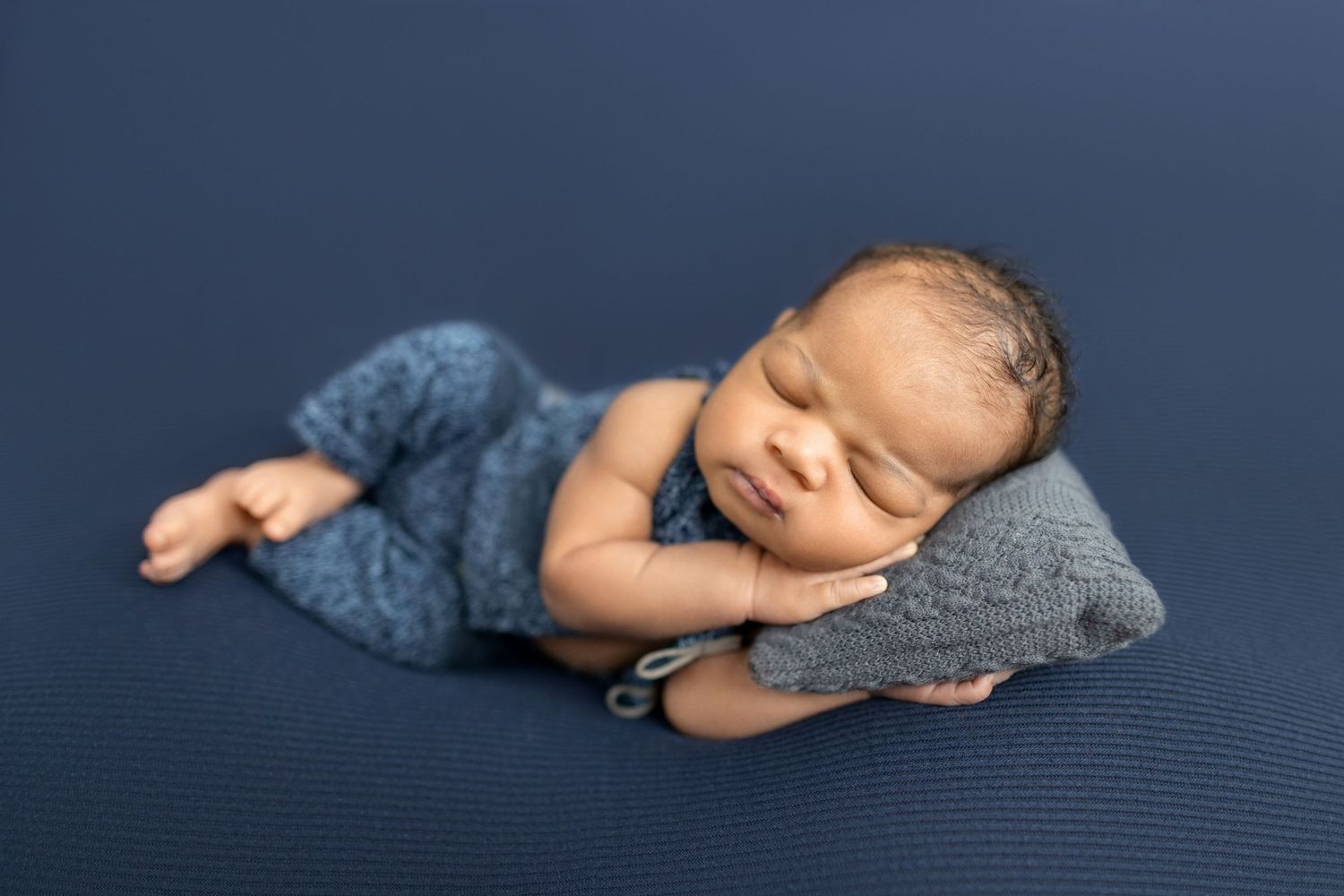 Sleeping newborn baby in blue knit outfit resting on tiny pillow on navy blue backdrop.