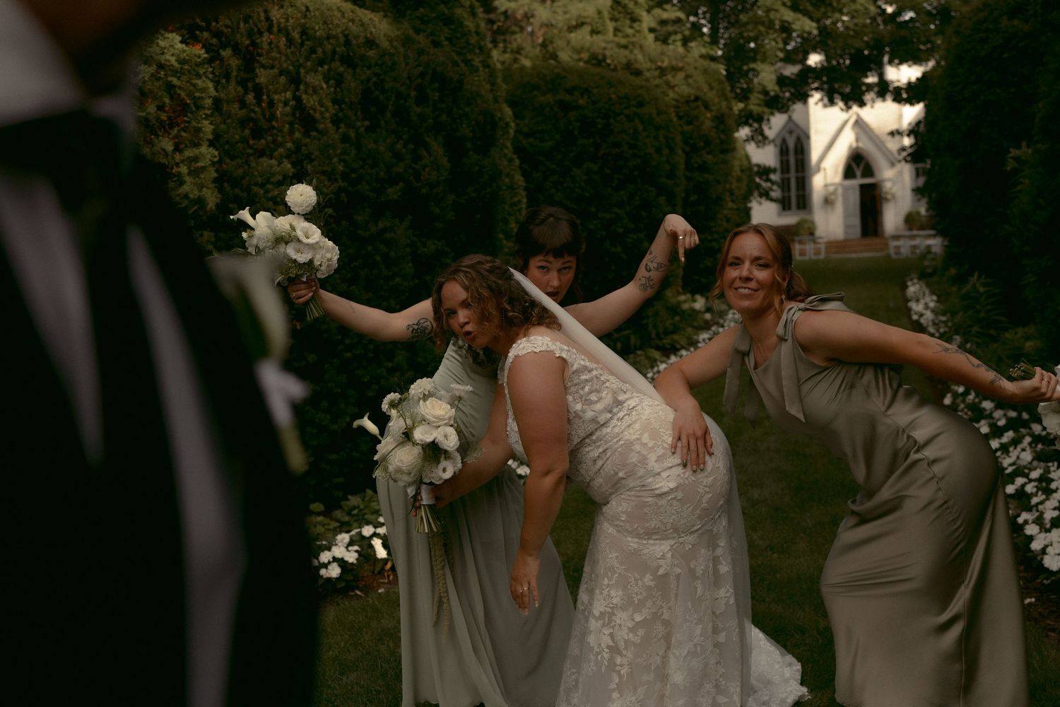 Bride in lace gown laughs with bridesmaids in sage dresses outdoors near a white chapel. Cranberry Creek Gardens Ontario