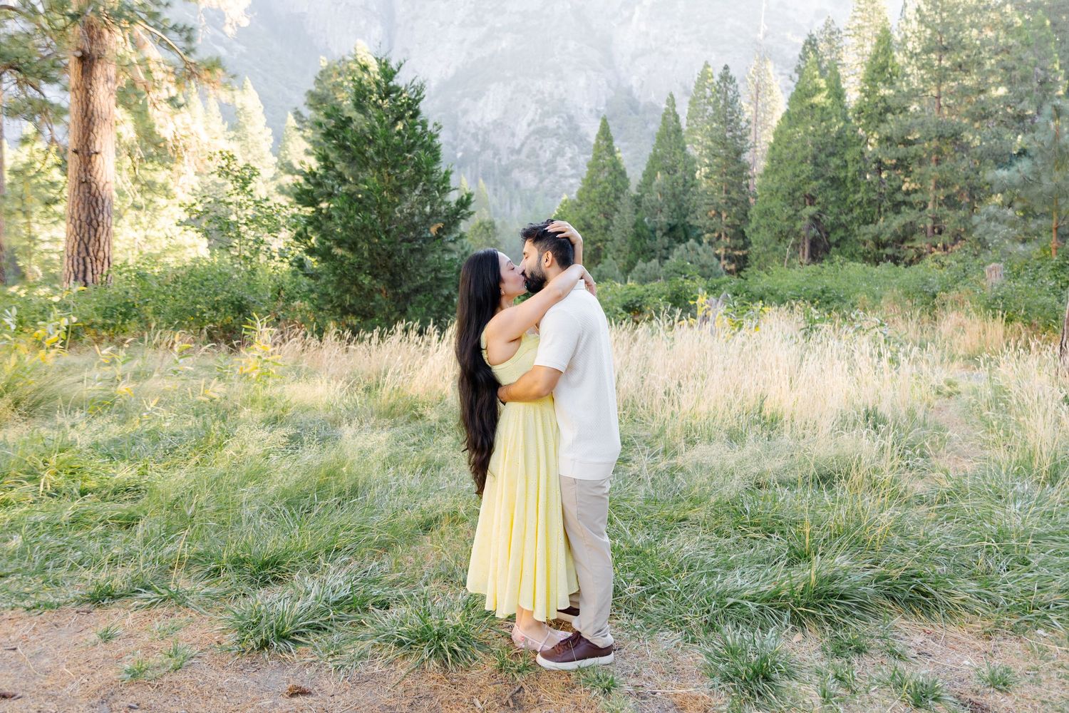 Couple sharing a romantic kiss in Yosemite meadow, woman in yellow dress, surrounded by tall pines and granite cliffs.