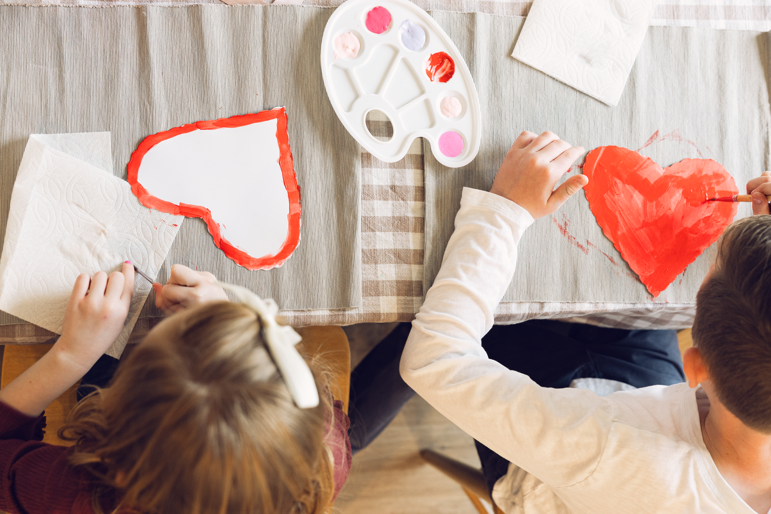 Two children painting red heart artwork together at table with watercolor palette overhead view.