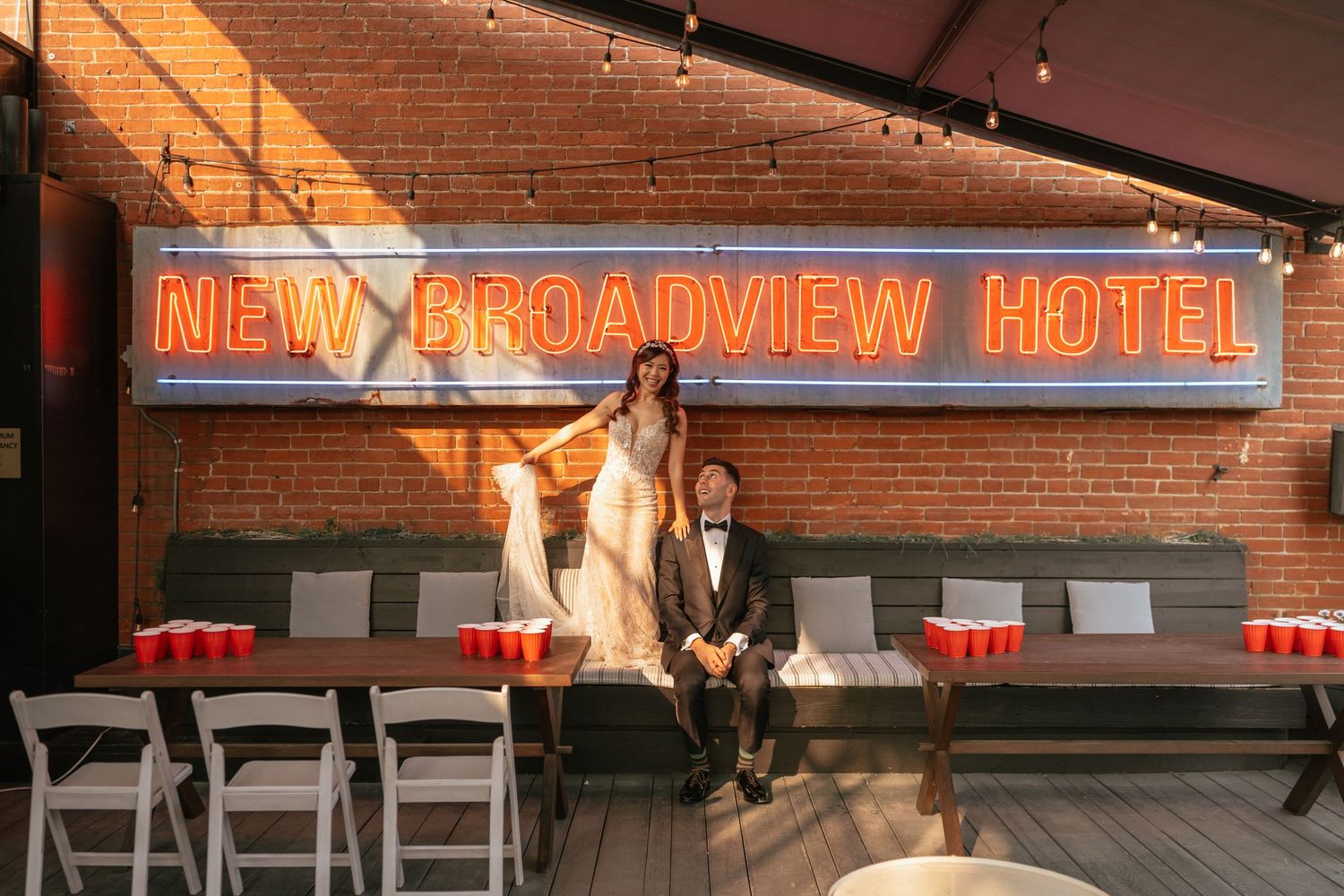 Couple posing in front of neon New Broadview Hotel sign on brick wall rooftop patio with string lights.