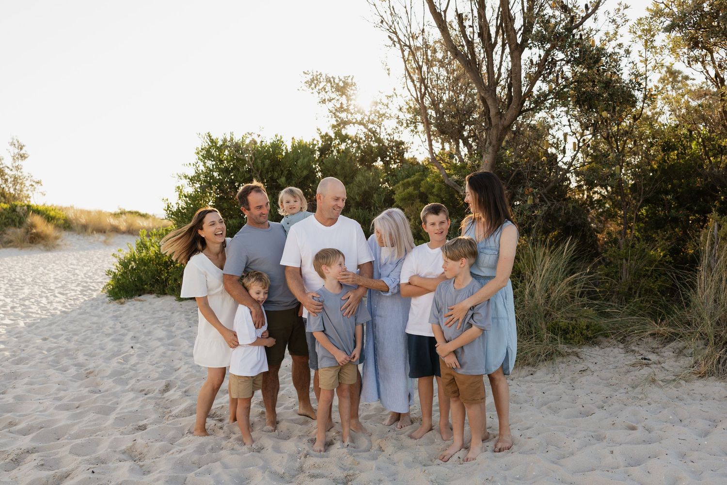Large family group photo on sandy beach with trees, wearing coordinated blue and white outfits in warm golden light.