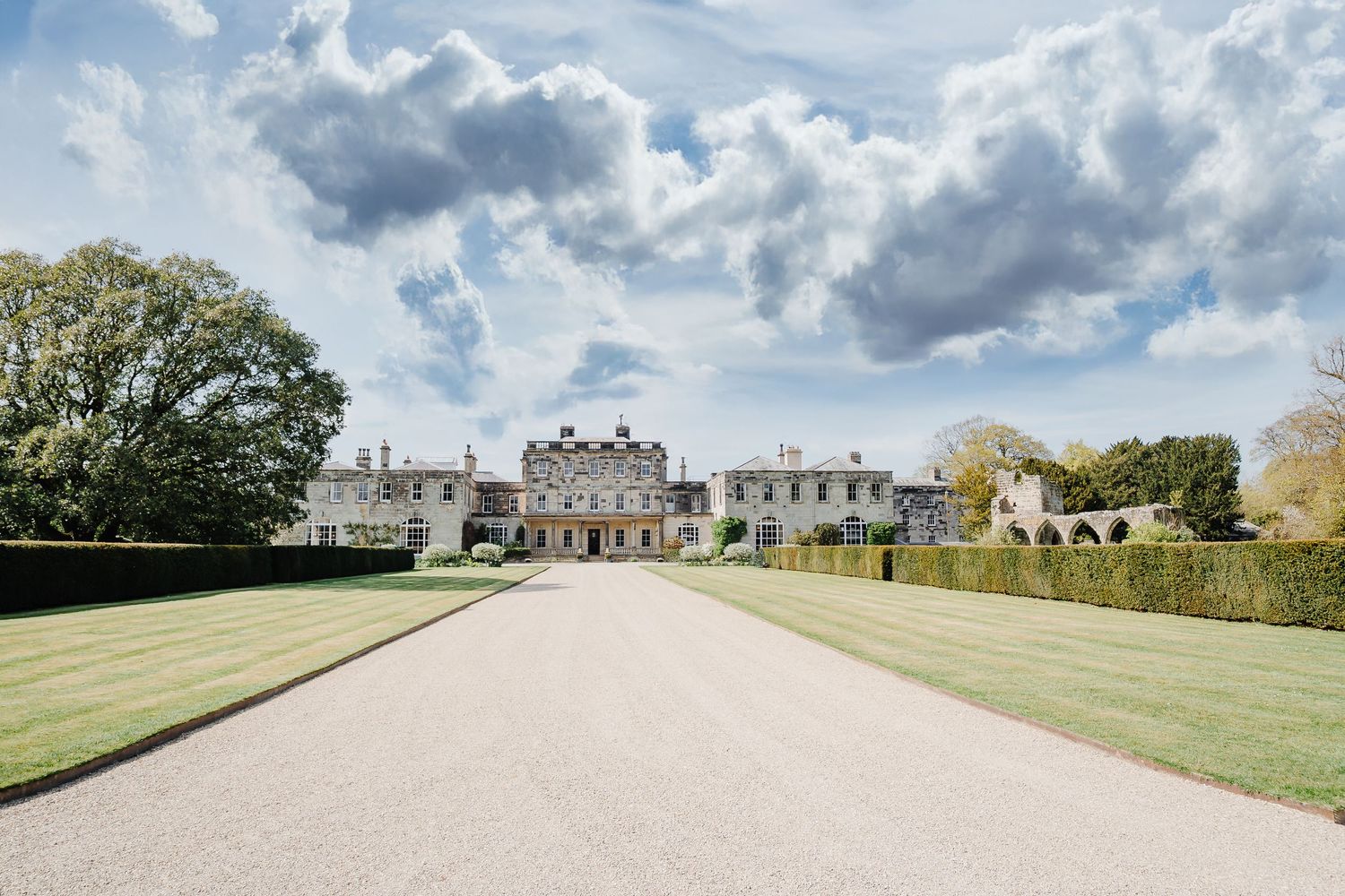 Grand English country estate with long gravel driveway, manicured hedges, and green lawns under dramatic cloudy sky.