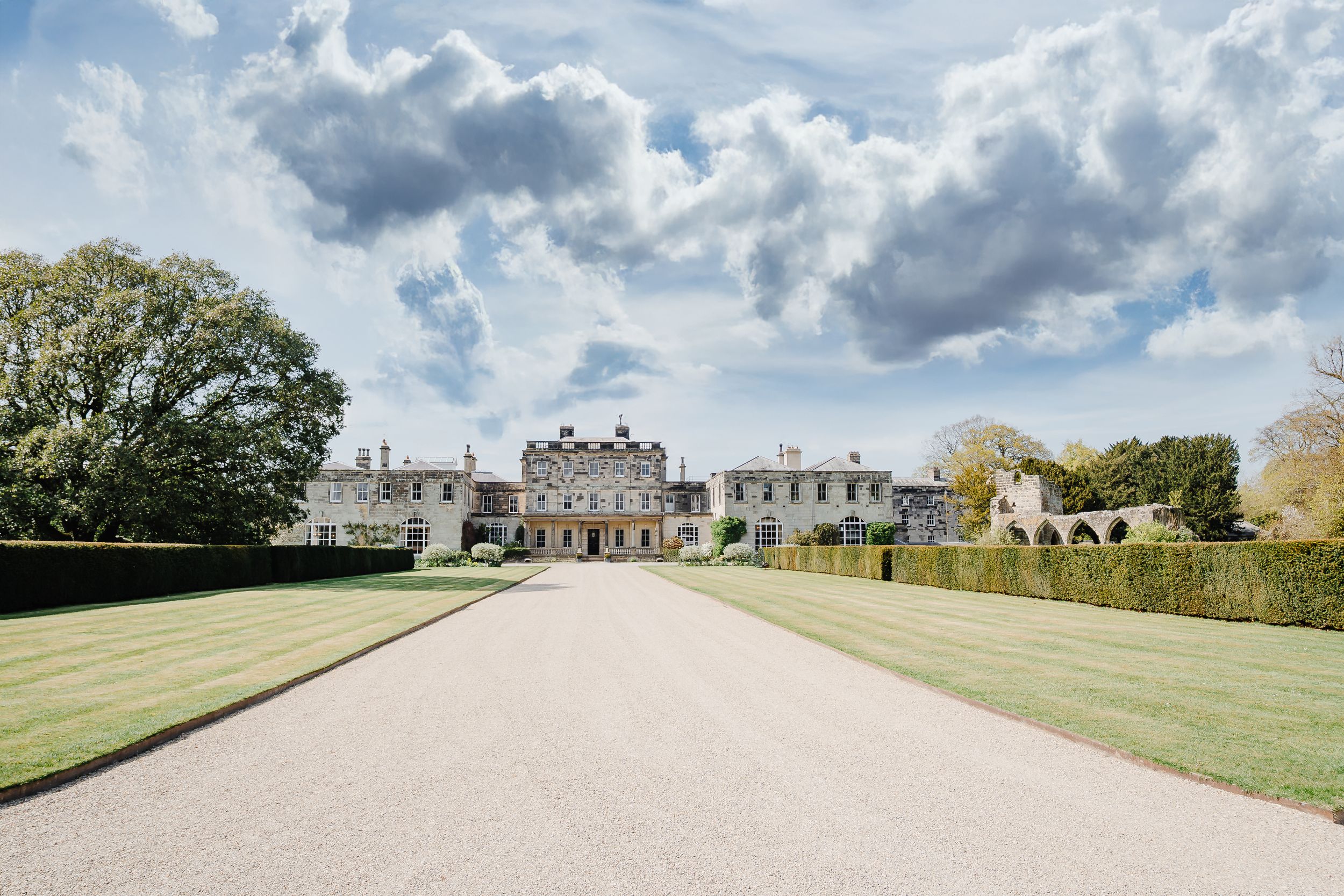 Grand English country estate with long gravel driveway, manicured hedges, and green lawns under dramatic cloudy sky.