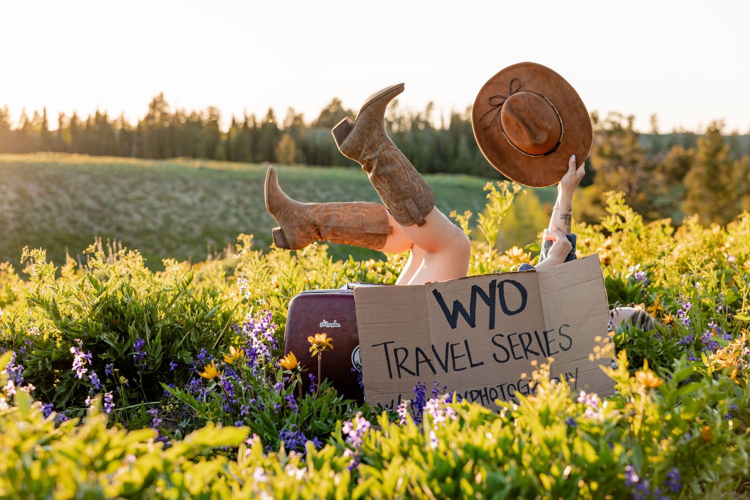 Person relaxing in wildflower field holding cowboy hat with WYO Travel Series sign and cowboy boots up in the air.
