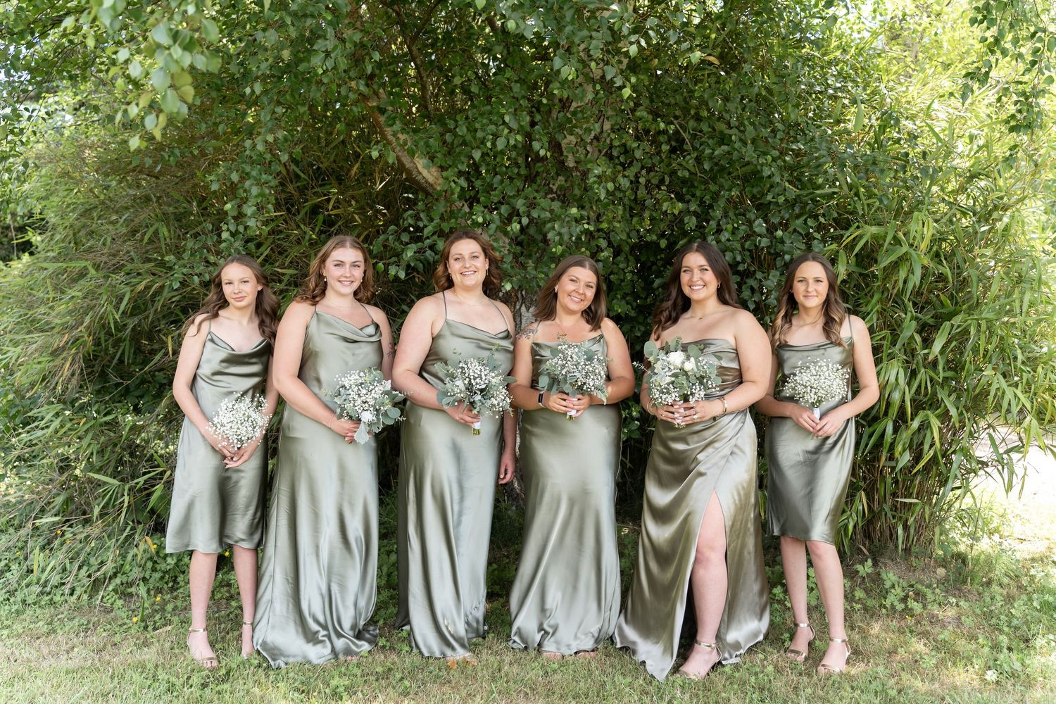 Six bridesmaids in sage green satin dresses holding white bouquets pose outdoors in front of lush green trees.