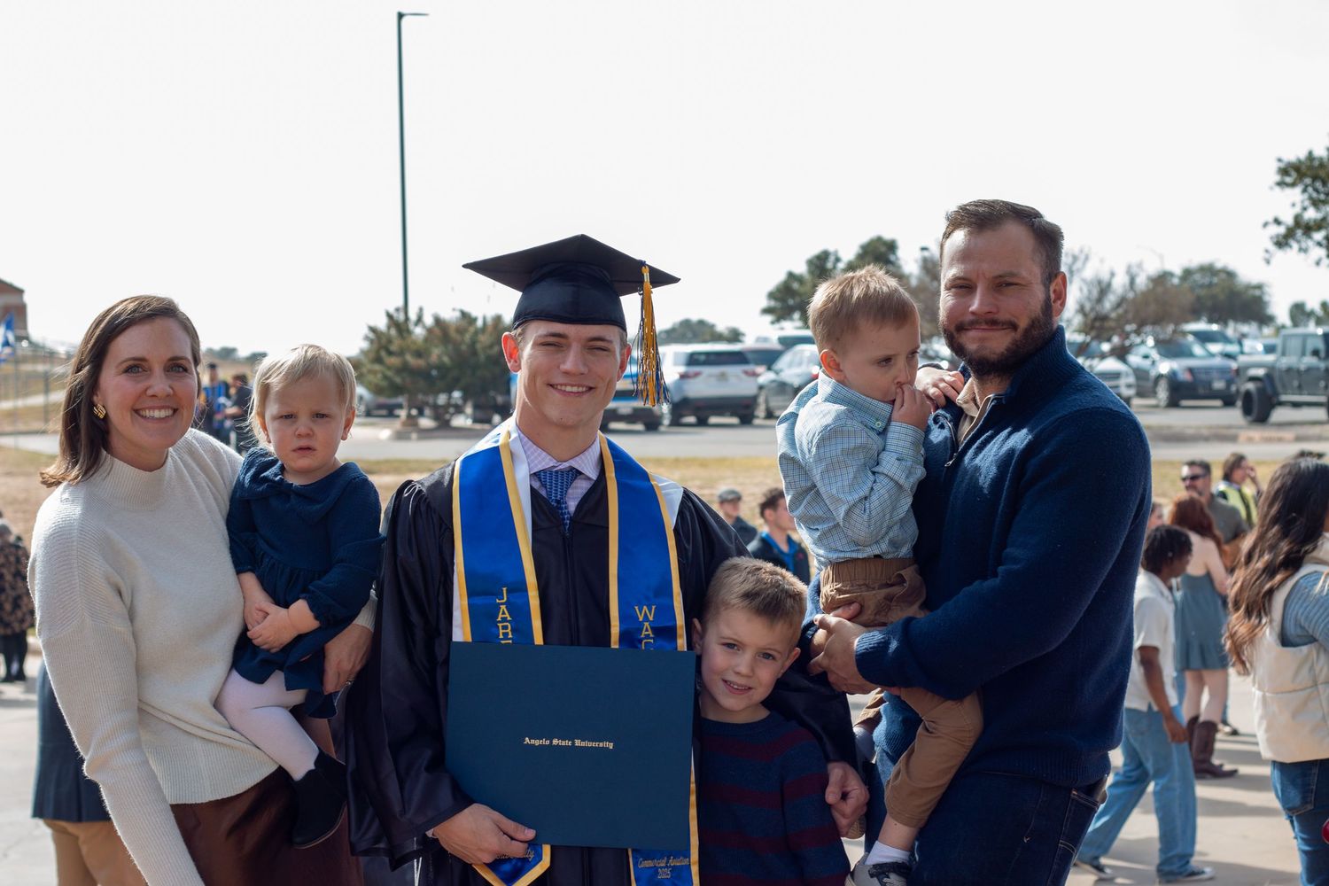 College graduate in cap and gown holds diploma surrounded by proud family outdoors at graduation ceremony.