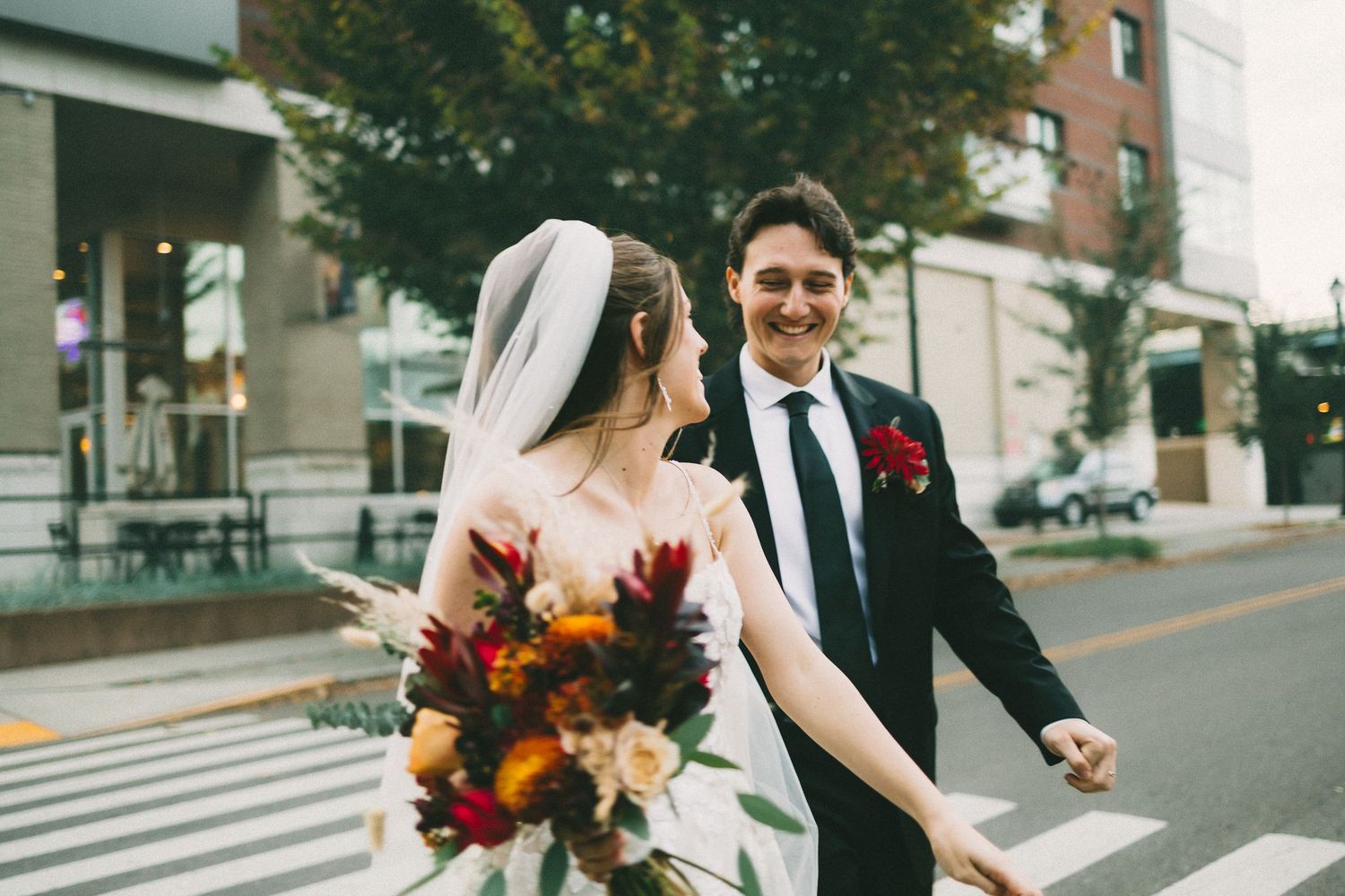 A romantic wedding couple shares tender moments along a tree-lined path with autumn leaves scattered on the ground.