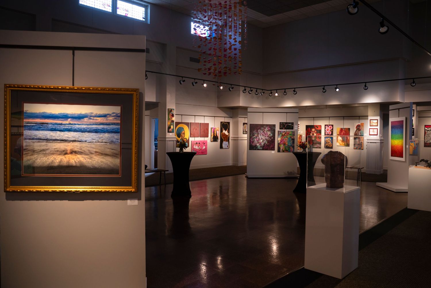 Art gallery interior showcasing colorful paintings and sculptures on display under warm lighting.
