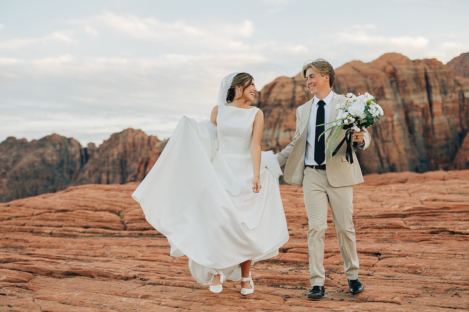 Bride in white gown and groom in tan suit hold hands on red rock desert landscape during outdoor wedding ceremony.