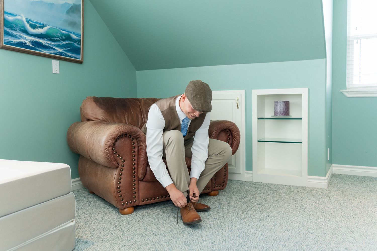 Man in vest and cap tying dress shoes while seated in brown leather armchair in teal attic room.