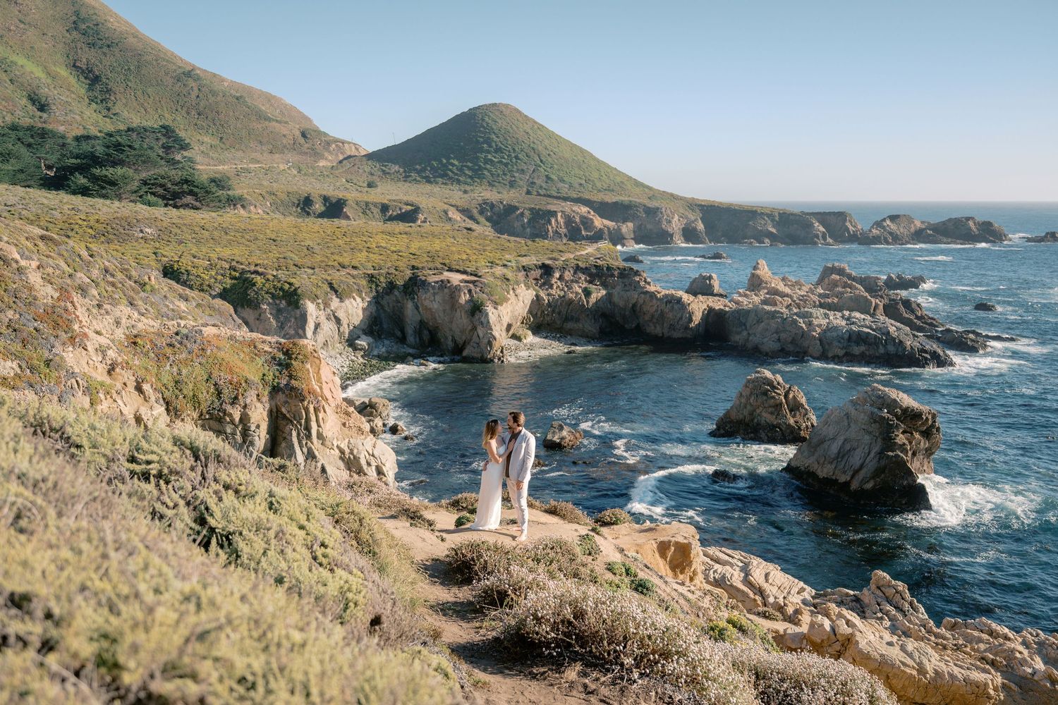 Couple standing on rocky Big Sur coastline with dramatic ocean views, sea stacks, and green hills under clear blue sky.