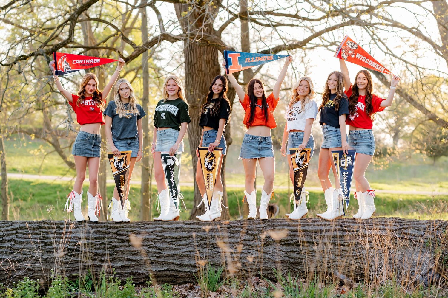Eight girls stand on a log holding college pennants outdoors, wearing university shirts and white boots.