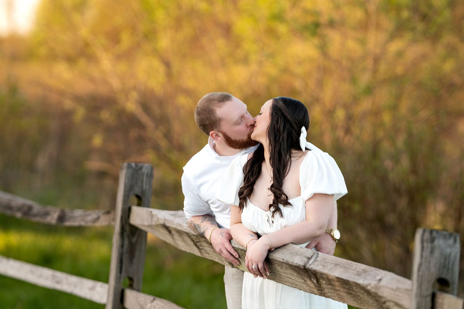 Couple sharing a romantic kiss leaning against a rustic wooden fence at golden hour.