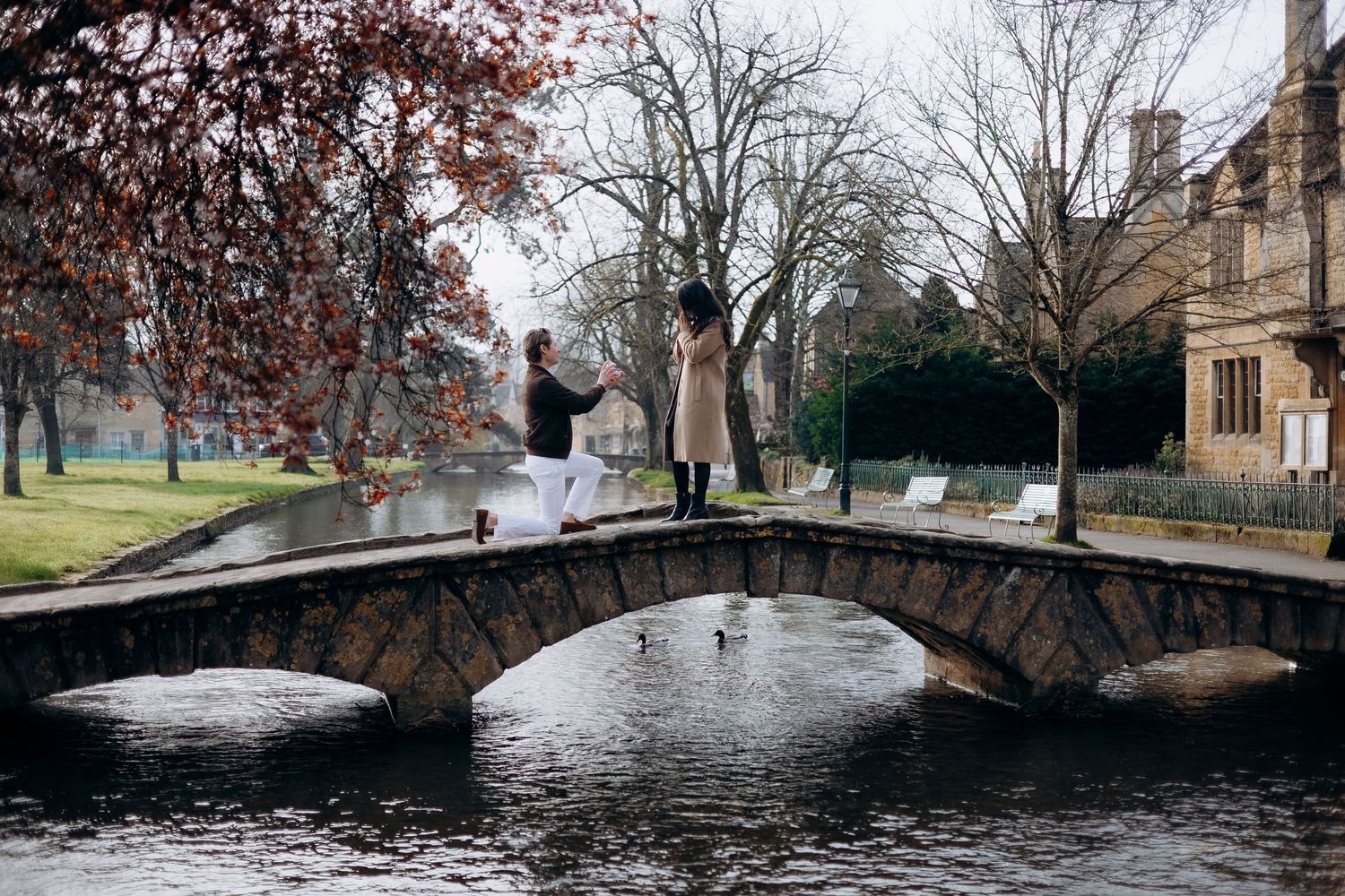 Couple holding hands on a stone bridge over a calm river in a scenic park with bare winter trees.