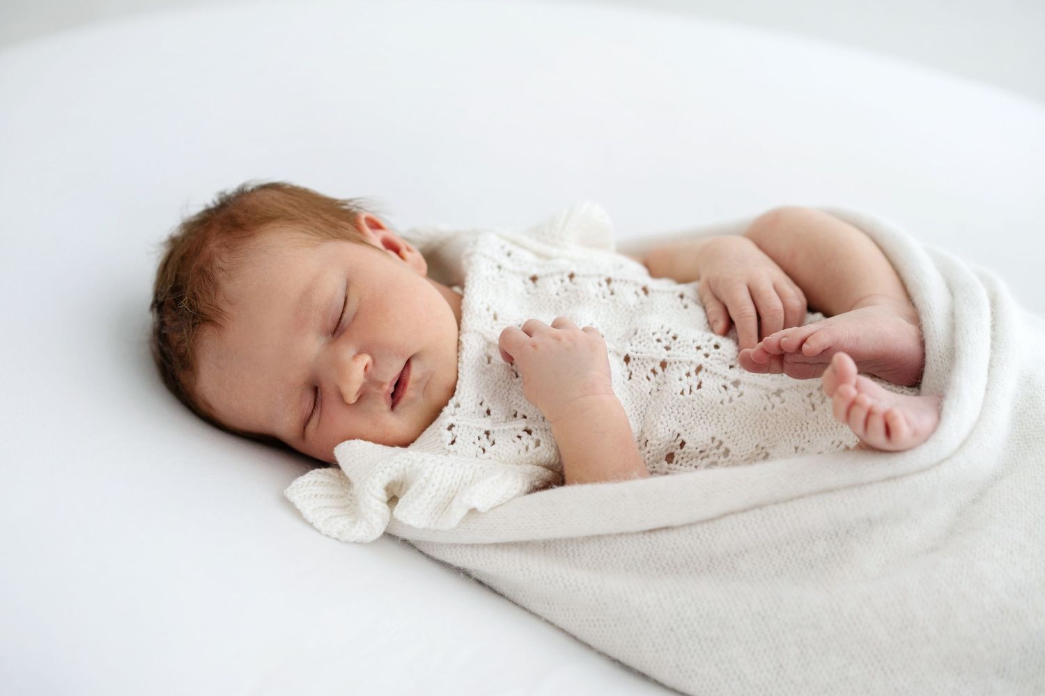 Peaceful newborn baby sleeping wrapped in a soft white knit blanket on a white background.
