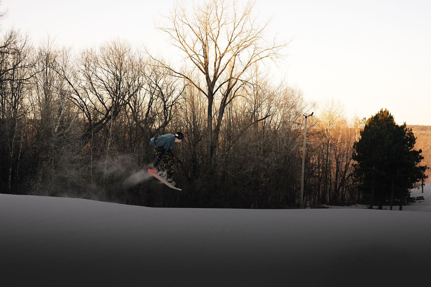 Snowboarder catching air on a snowy slope at sunset with bare winter trees in background.