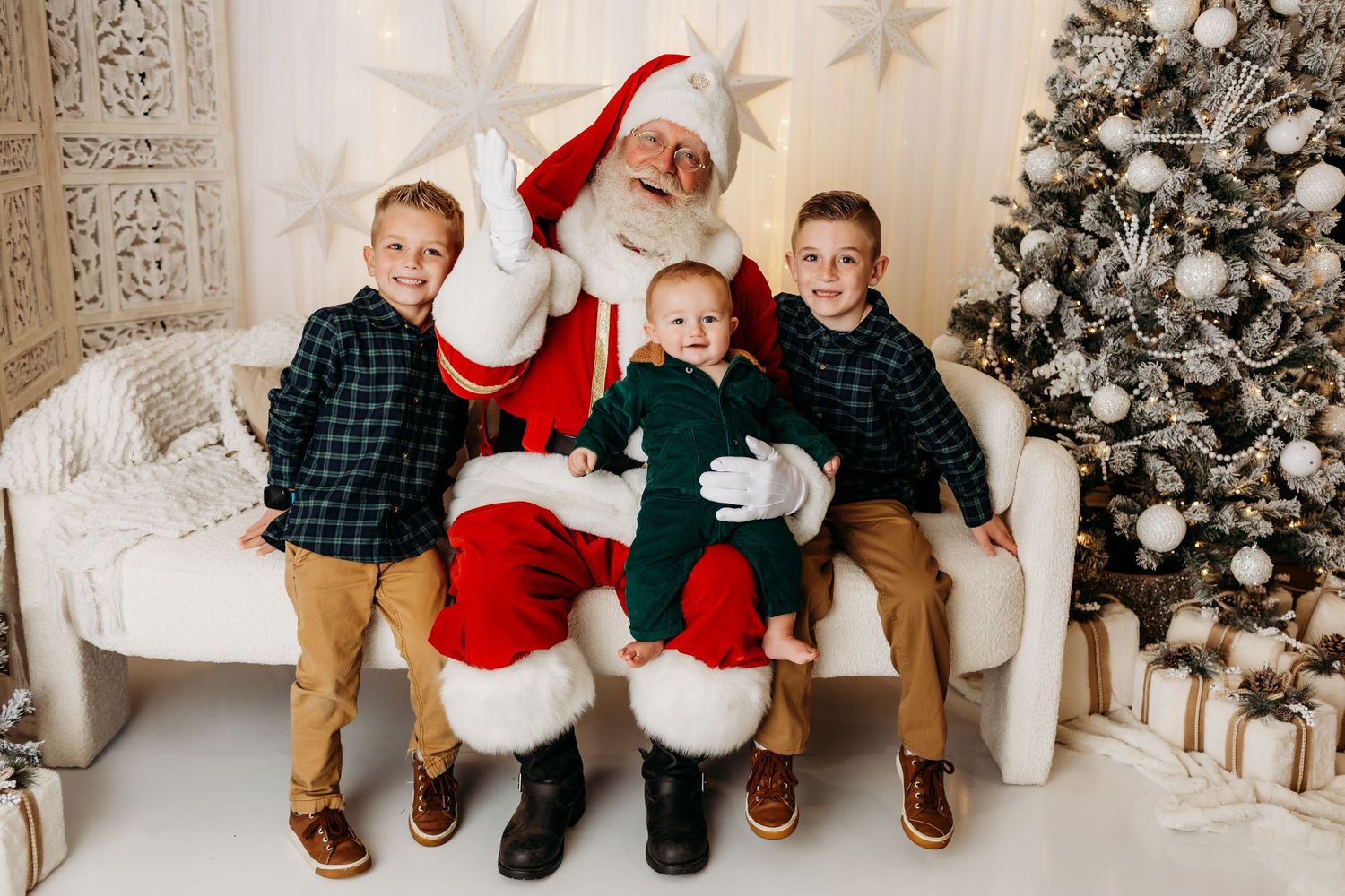 Three boys pose with Santa Claus on a white bench beside a decorated Christmas tree with gifts.