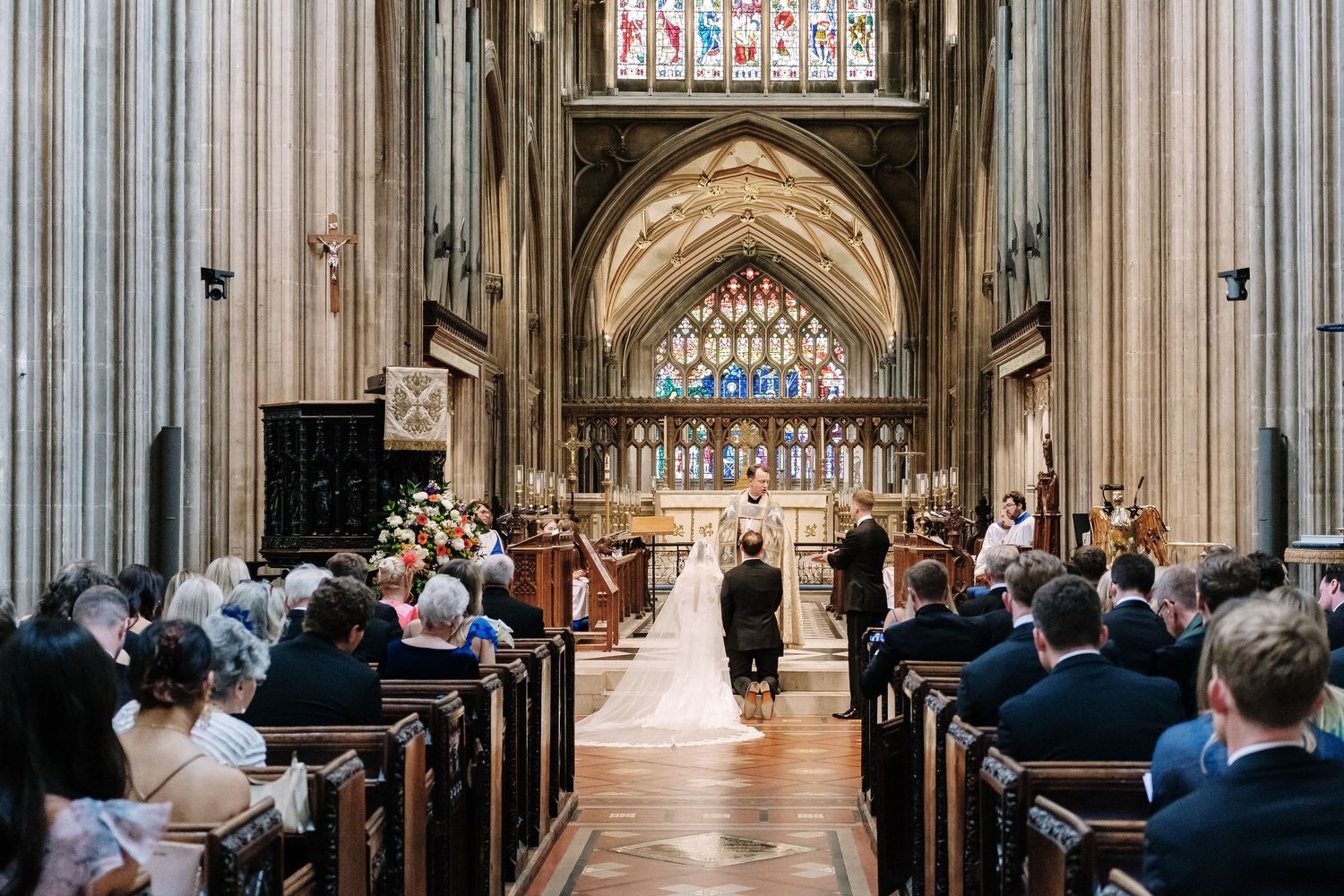 Wedding ceremony in St Mary Redcliffe Church with stained glass windows, guests seated in wooden pews, bride and groom at the altar.