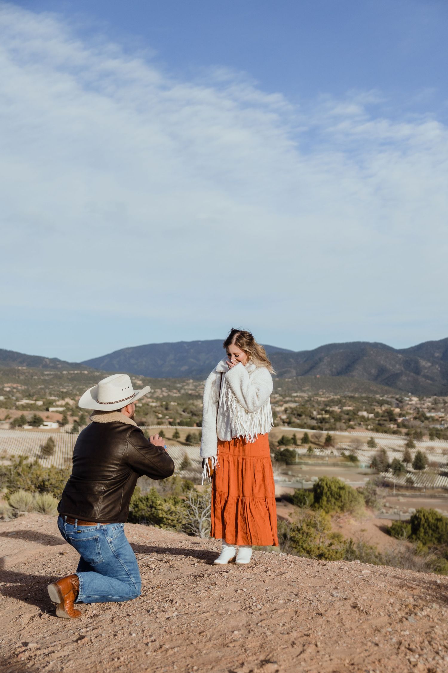 Man on one knee proposing to woman in orange skirt and white jacket with mountain landscape backdrop.