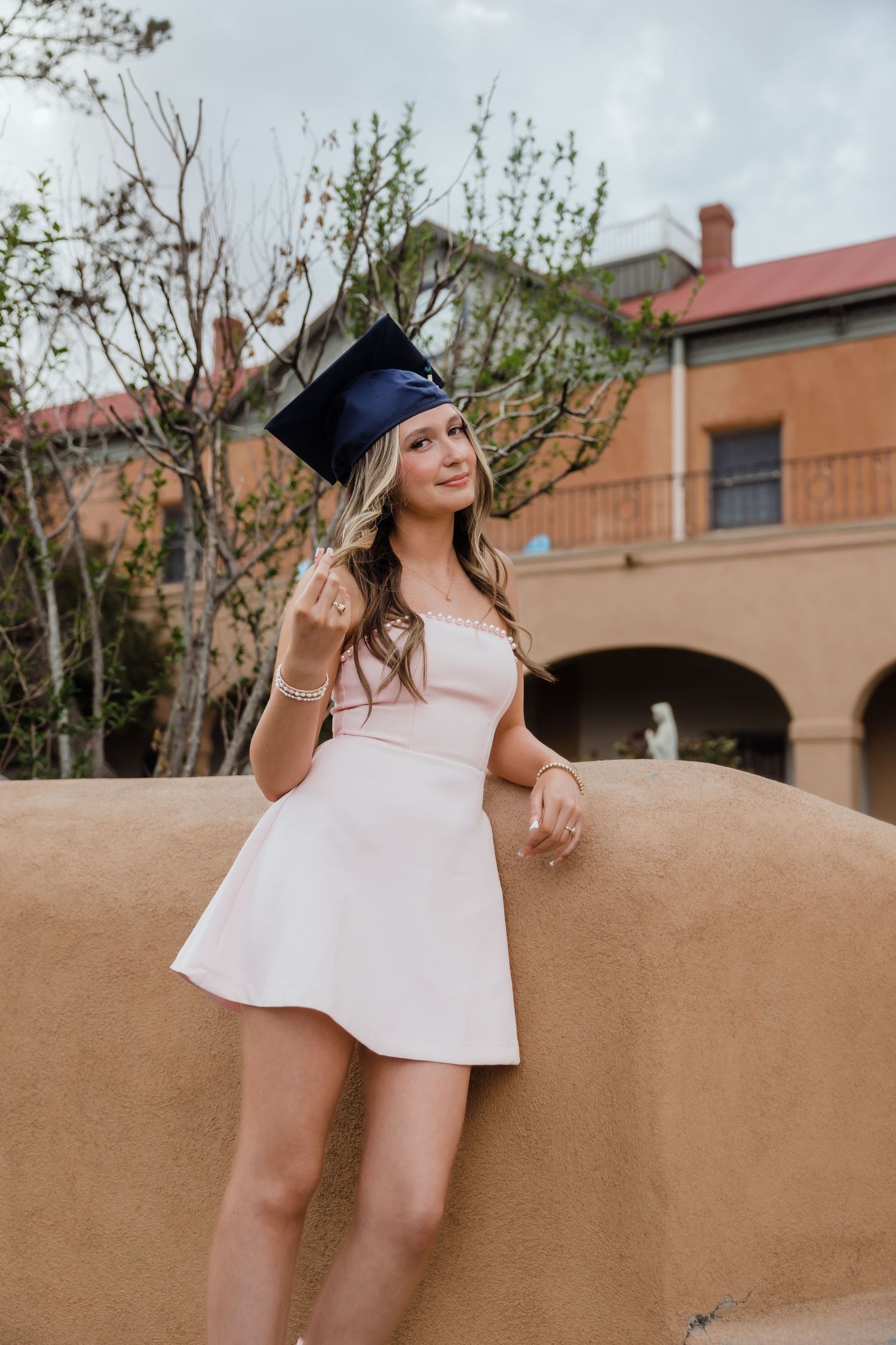 Young woman in white mini dress and navy graduation cap poses outdoors near adobe wall, celebrating her graduation.