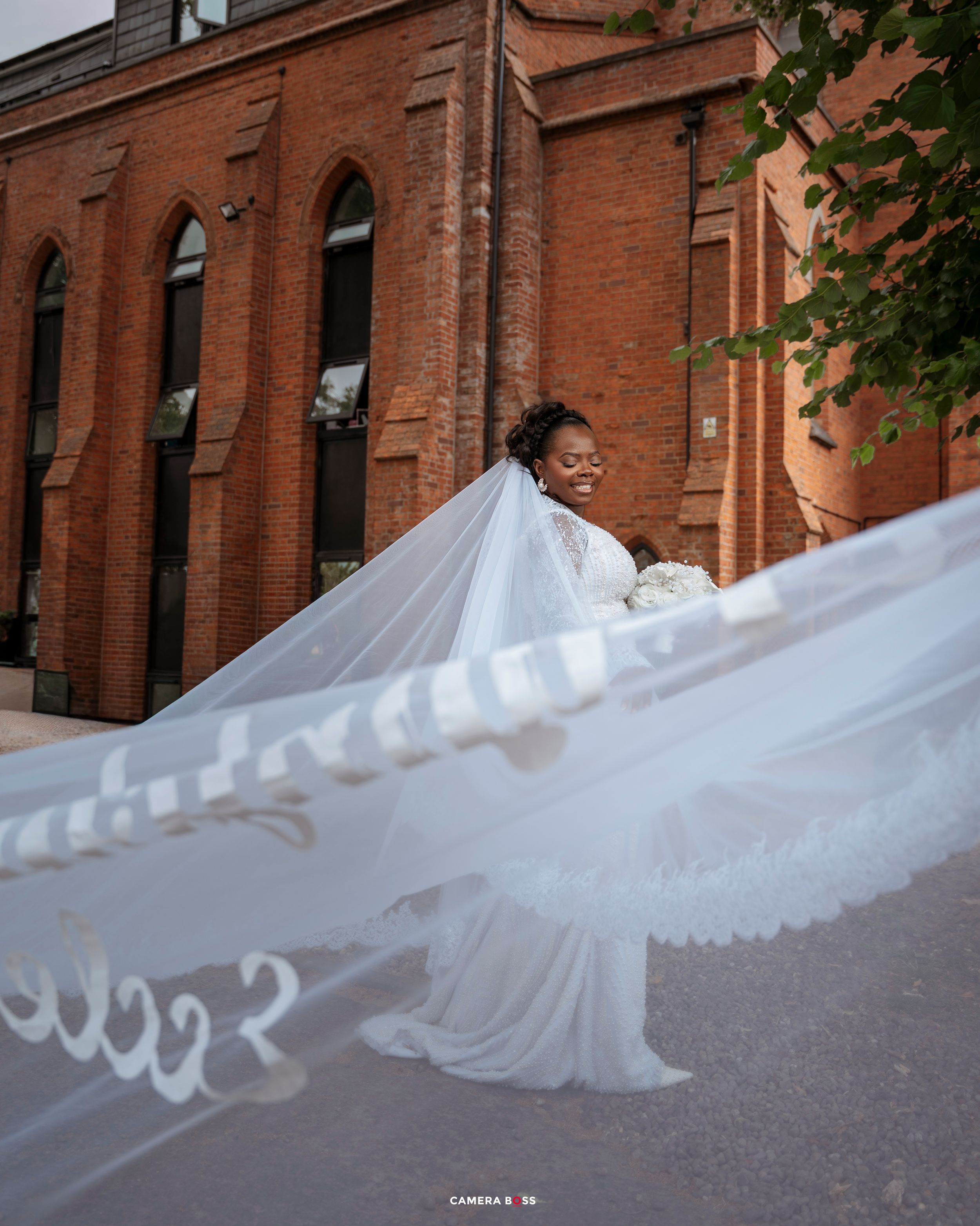 Bride in white gown with flowing veil billowing in wind outside red brick church building.