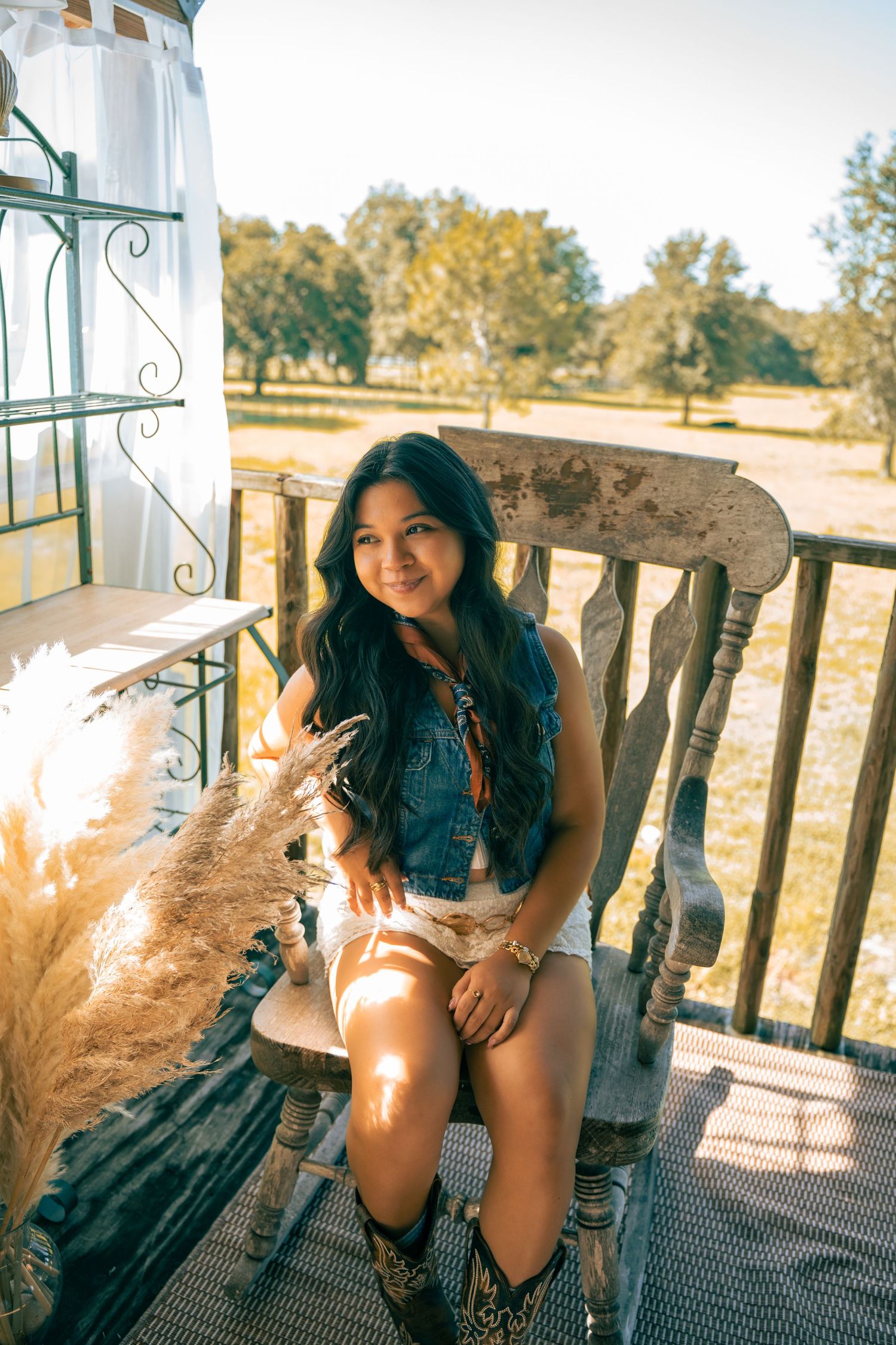 Smiling young woman in denim vest sits on rustic wooden chair on sunny porch with pampas grass decor.