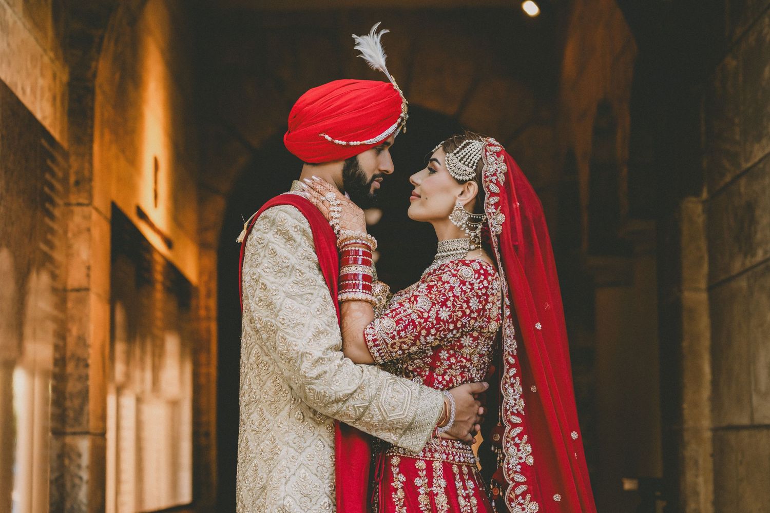 Indian bride and groom in traditional red wedding attire share a romantic moment in an ornate archway.