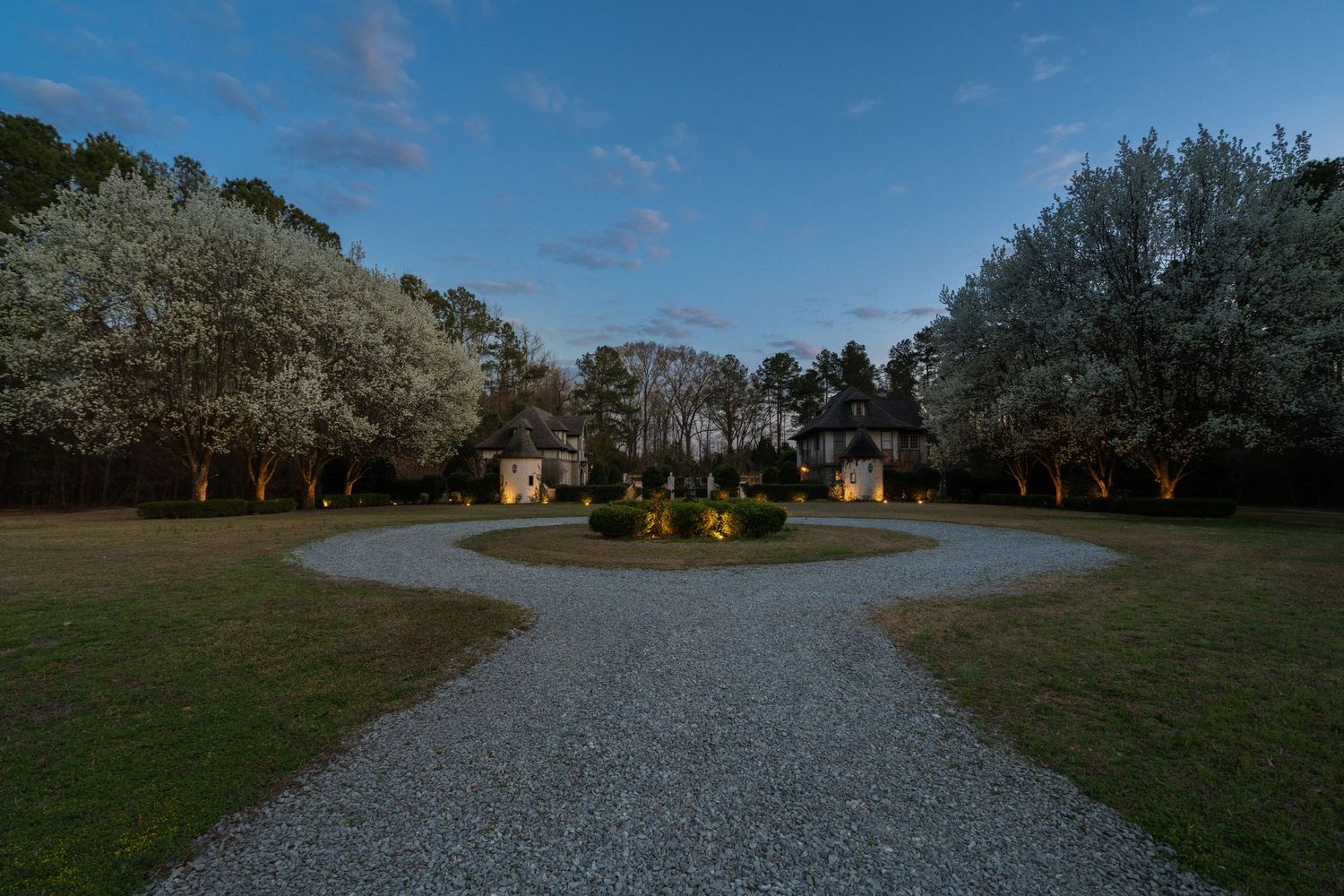 Circular gravel driveway with blooming white trees flanking a grand estate at dusk with soft landscape lighting.