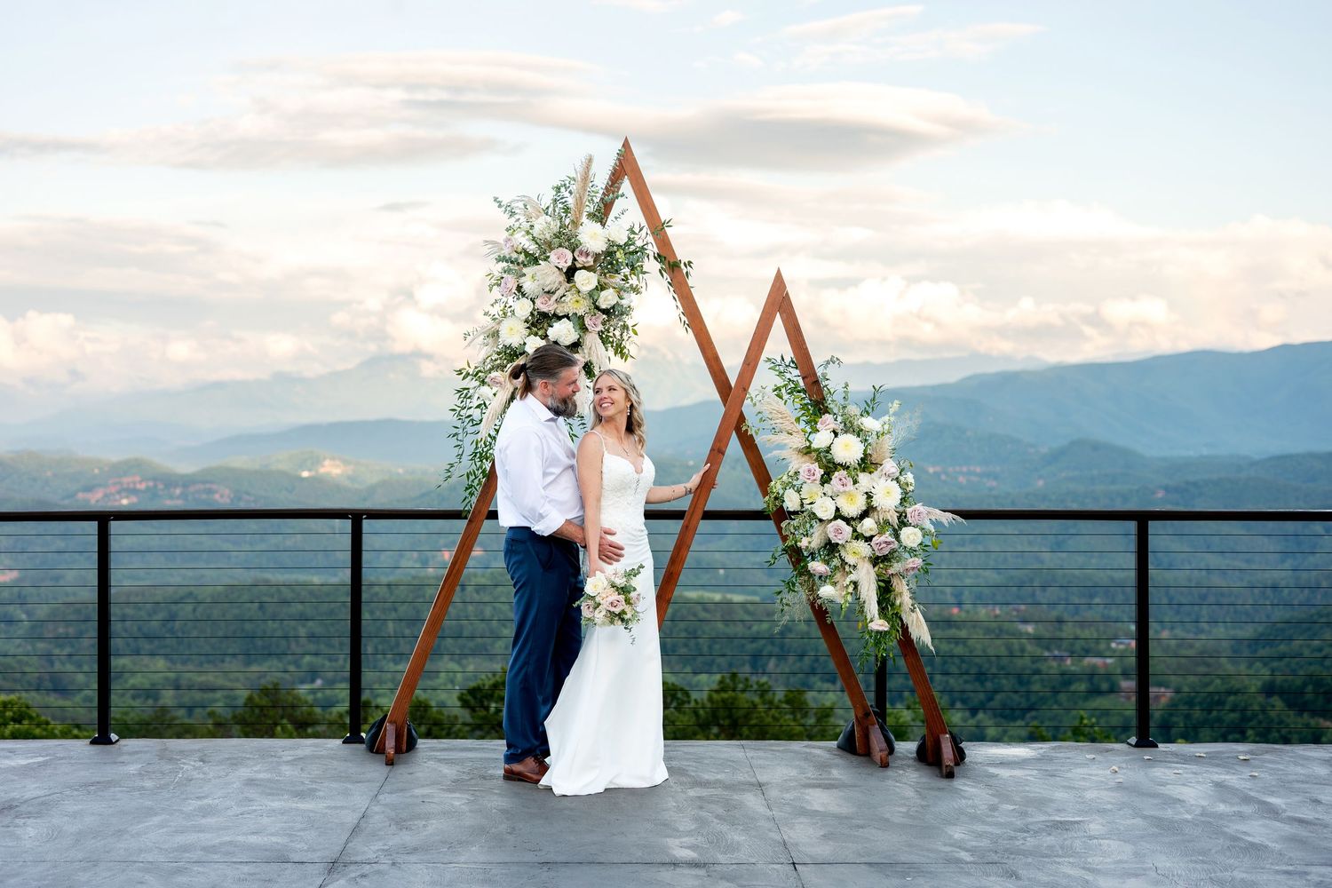 Couple embraces under a triangular wooden arch adorned with white florals, with mountain views at an outdoor wedding ceremony.