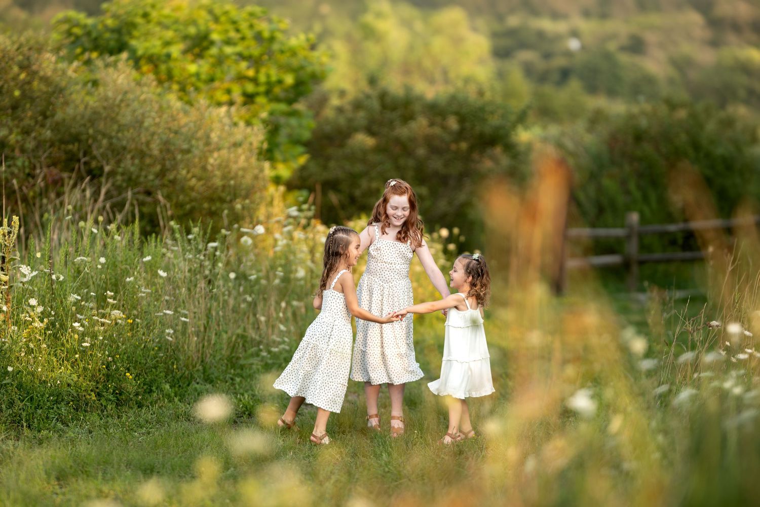 Three girls in white dresses hold hands in a sunlit meadow surrounded by wildflowers and lush greenery.
