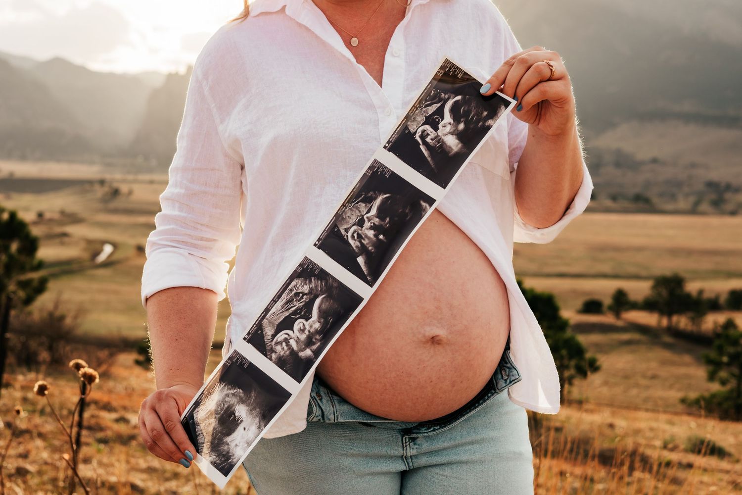 Pregnant woman outdoors holding ultrasound strip photos showing her baby bump in mountain landscape| Maternity Photos in Boulder Colorado