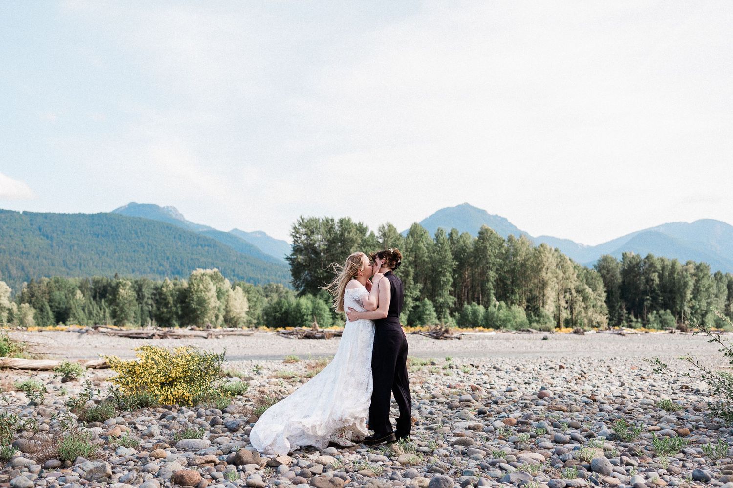 two brides kiss on a windy day in front of the Cowltiz River during their Packwood elopement in Washington.