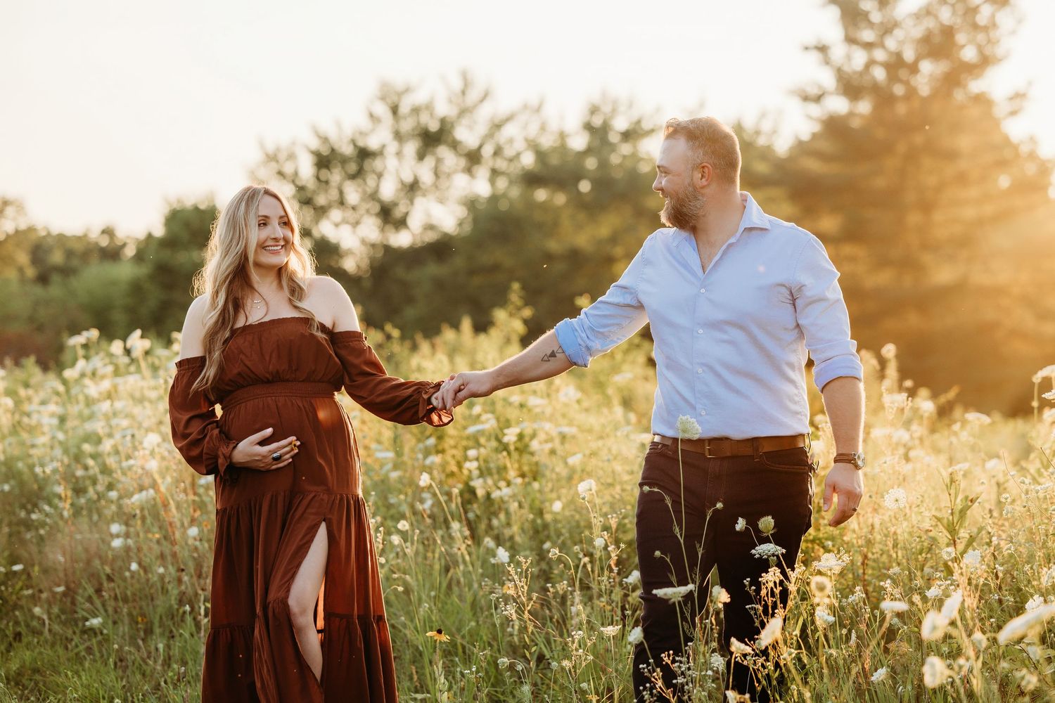 Pregnant couple holding hands in a sunlit wildflower field, woman in rust dress cradling baby bump.